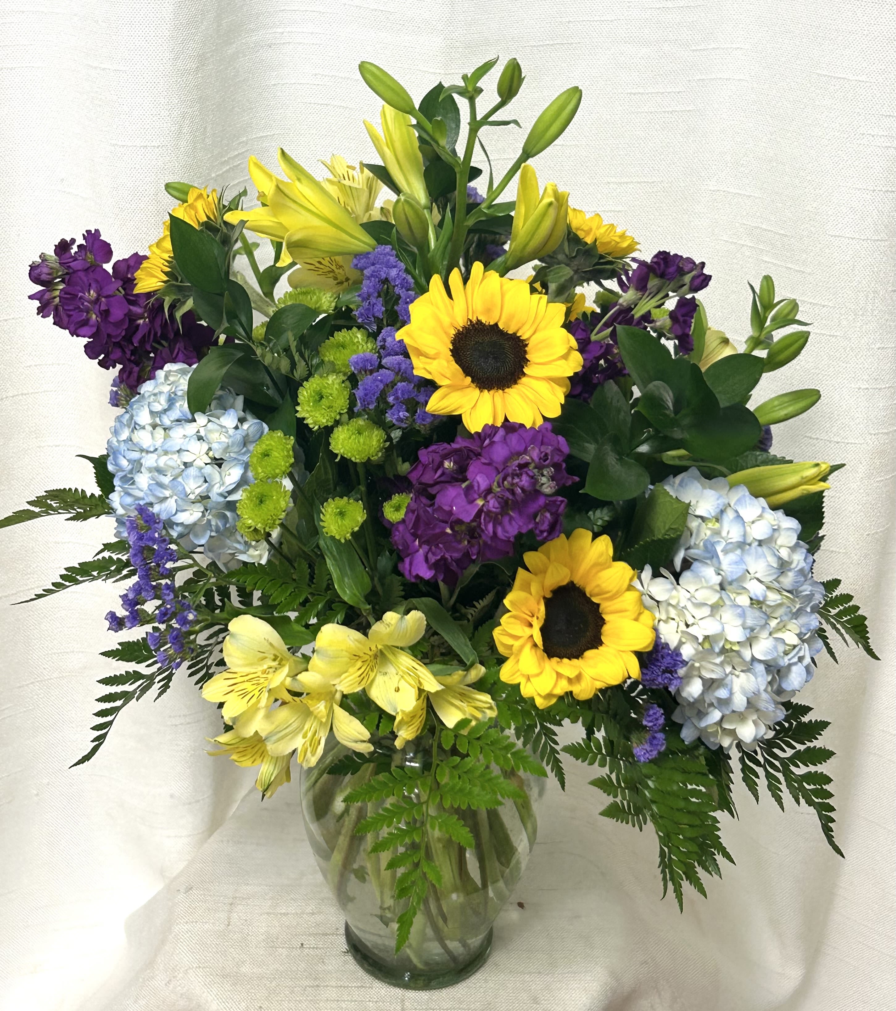 Summer Breeze (BB) - Experience the essence of sunshine and warmth captured in our &quot;Summer Breeze&quot; floral arrangement, a delightful composition that embodies the carefree spirit and vibrant colors of the summer season. This enchanting ensemble features the iconic radiance of sunflowers, the cool serenity of blue hydrangea, the velvety richness of purple stock, the sunny charm of yellow alstroemeria, and the golden elegance of yellow lilies, creating a harmonious blend of hues and blooms that evoke the joy and beauty of lazy summer days.