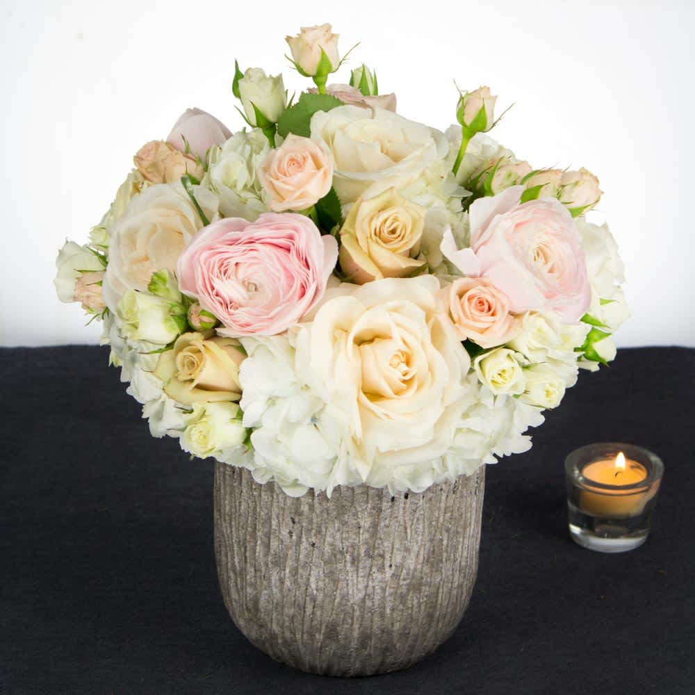 Made Me Blush (BB) - Our funeral arrangement is a beautiful and comforting display, thoughtfully designed to honor and celebrate the life of your loved one. This arrangement features a delicate blend of cream, white, and pink flowers, including roses, hydrangea, and mini spray roses, elegantly presented in a ceramic or stone container.