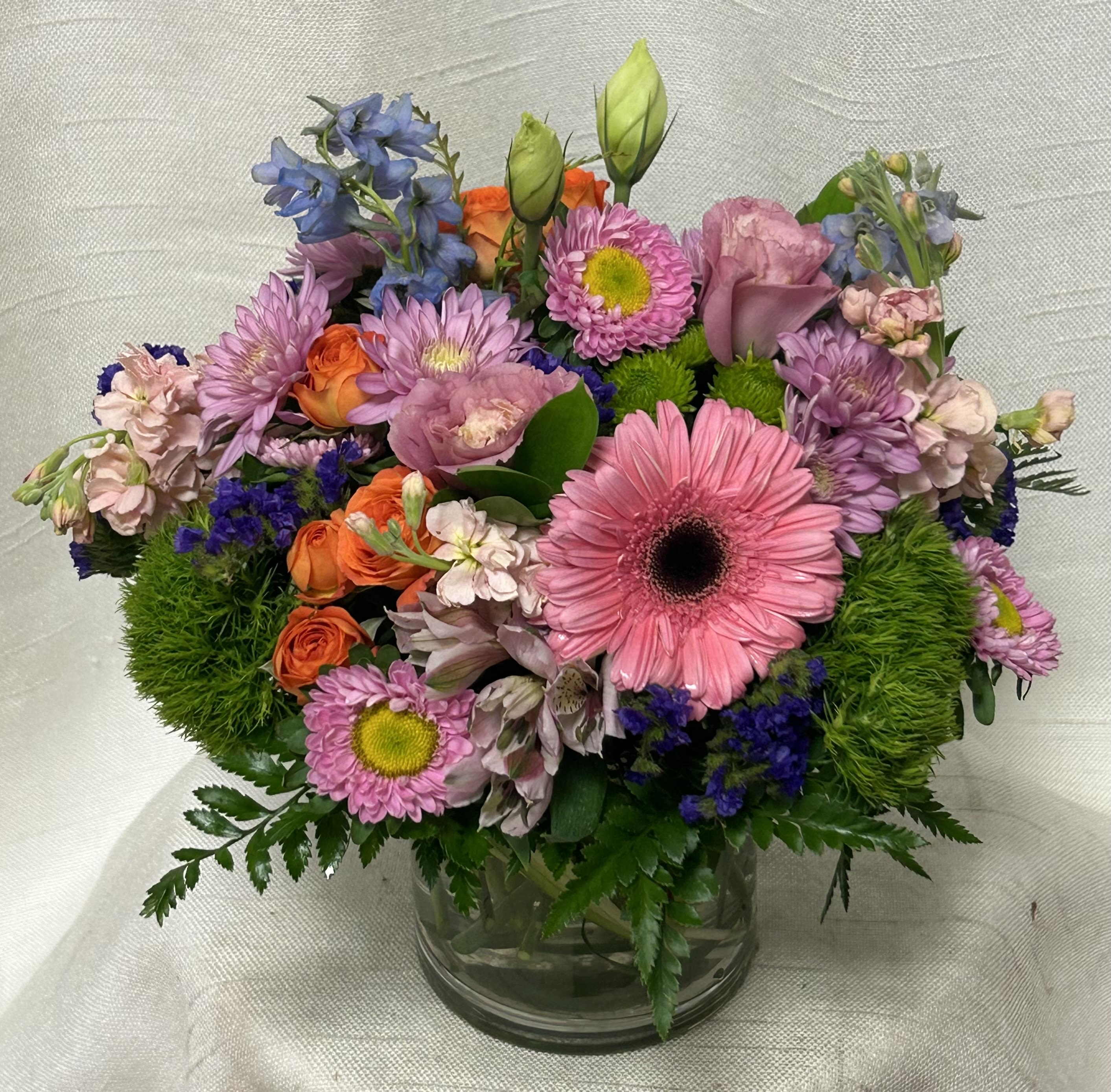 Enamored (BB) - &quot;Enamored&quot; is a captivating and vibrant flower arrangement that exudes joy and romance. Bursting with a kaleidoscope of hues, this enchanting bouquet is a harmonious blend of pink gerbera daisies, delicate pink lisianthus, and cheerful orange spray roses. The addition of fragrant pink stock and the regal presence of blue delphinium elevate the arrangement, adding depth and contrast.