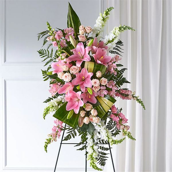 Elegant Embrace (BB) - Introducing our Elegant Embrace Standing Spray, a heartfelt and elegant expression of love and remembrance. This exquisite arrangement features pink lilies, pink spray roses, white snapdragons, and lush greens, all beautifully arranged to create a stunning display that pays homage to your loved one.