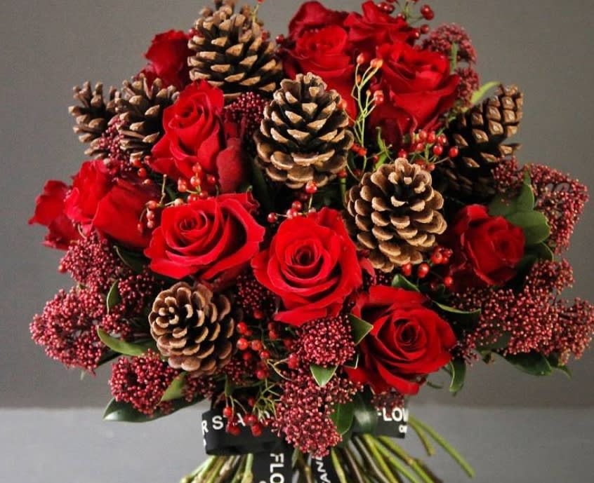 Spice Spice - A beautiful handheld design using red roses and pine cones with Christmas greens. Bouquet will be filled to value depending on which size is chosen. Premium version shown.