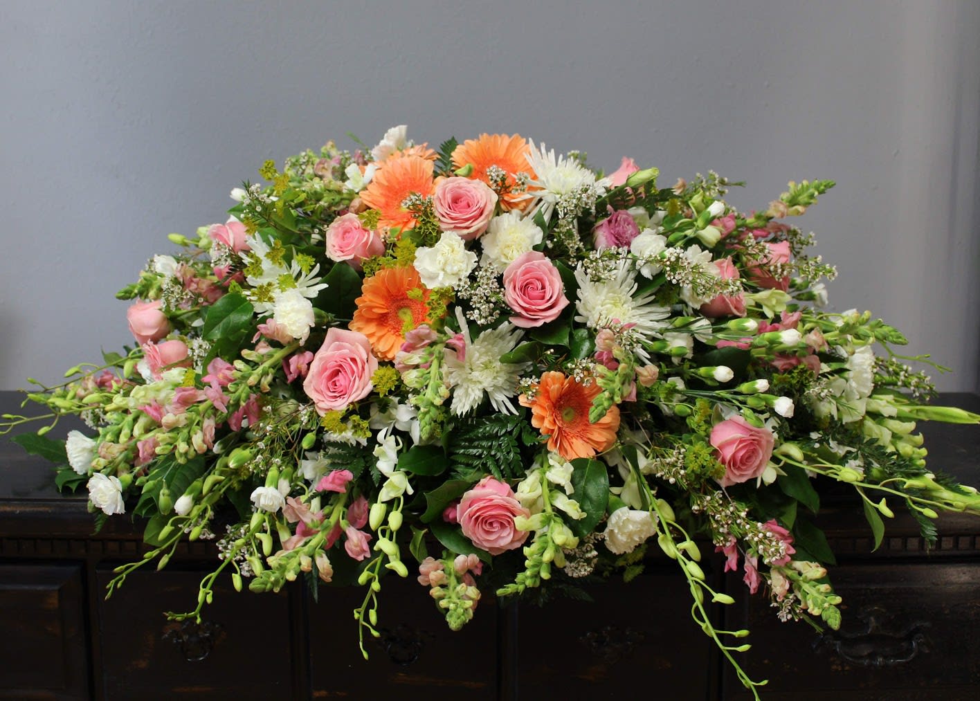 FUNERAL CASKET SPRAY - Warmest Memories 82 - This is a wonderful option for a funeral service or church. This casket can be designed with a saddle that rests over approximately half of the casket for an open casket or the full casket for a closed casket service. This lovely mix of peaches, pinks and white roses, daisies, snapdragons and orchids is a great way to add some gentle color to your service.   **This is a piece for a funeral or memorial service. Please place the order at least 2 days prior to the service to guarantee a certain arrangement. Anything sooner and we cannot guarantee it will match the picture. These pieces can be customized to the color of your choice. The flowers may vary depending on availability of the flowers, special requests and quality of the flowers we have in stock. If you have a special request please add it to our special instructions when placing your order. If you have any questions please don’t hesitate to call. 714 289 1123**