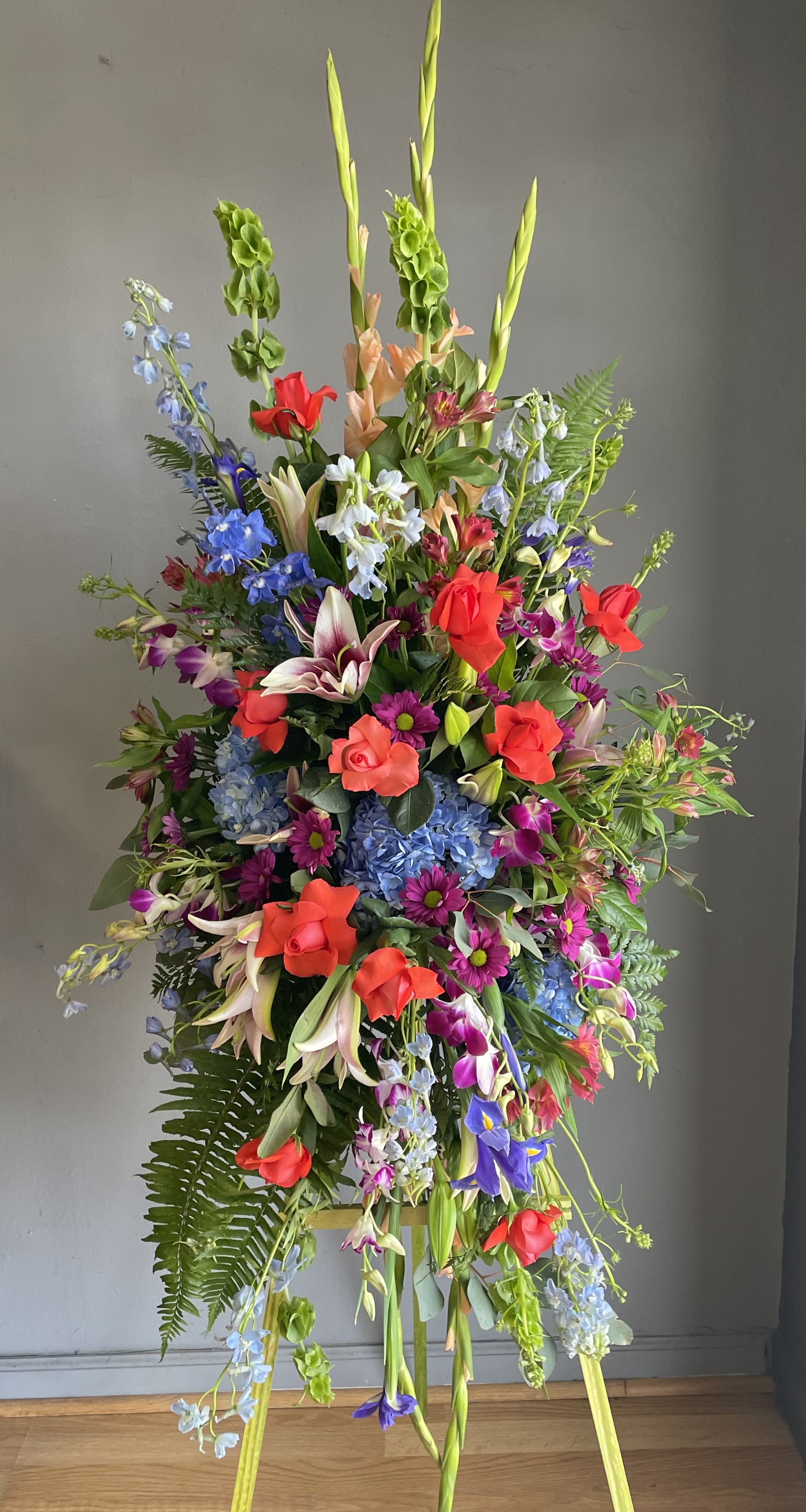 FUNERAL STANDING SPRAY - Garden Vibrancy 102 - This is a beautiful option for a funeral service, church or graveside. This standing spray is a one sided flower arrangement on a 5’ easel (easel included) displaying a spray approximately 4’ tall and 2’5” wide. This lovely mix of purple, orange and blue flowers garden flowers will add both a gentle and colorful touch to any day of memorial.   **This is a piece for a funeral or memorial service. Please place the order at least 2 days prior to the service to guarantee a certain arrangement. Anything sooner and we cannot guarantee it will match the picture. These pieces can be customized to the color of your choice. The flowers may vary depending on availability of the flowers, special requests and quality of the flowers we have in stock. If you have a special request please add it to our special instructions when placing your order. If you have any questions please don’t hesitate to call. 714 289 1123**