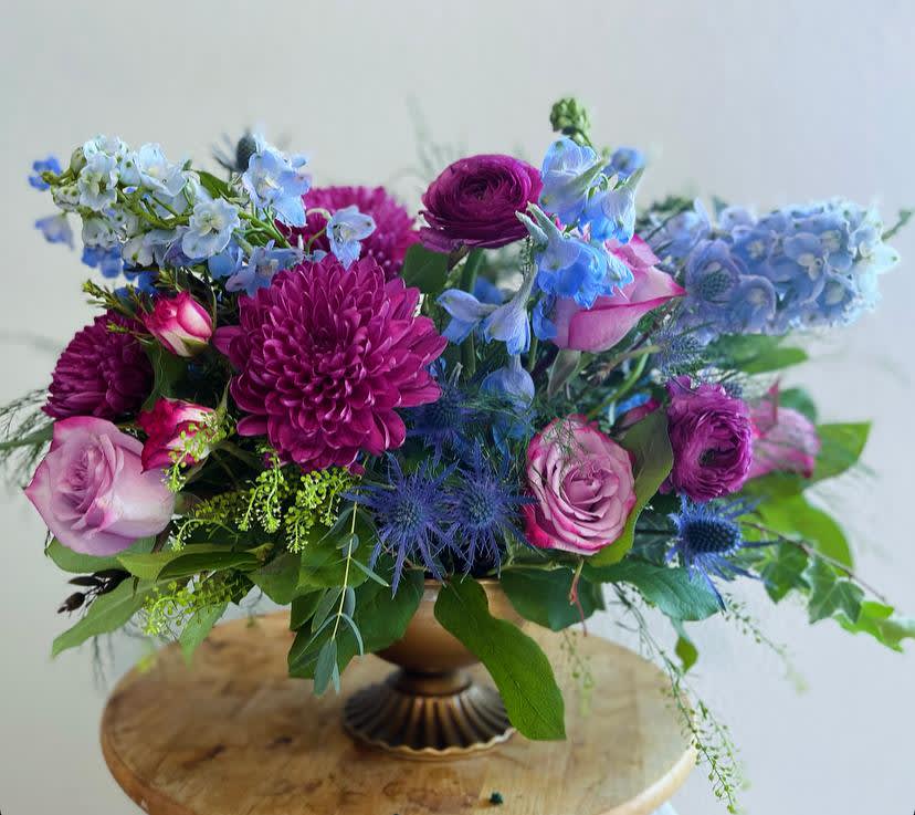 Chef's Kiss - Immerse yourself in the captivating beauty of our April May Flowers original cult favorite and top-selling arrangement, the Chef’s Kiss. This stunning floral masterpiece showcases a harmonious blend of moody gem tones that evoke a sense of mystery and allure. Lush blue delphinium cascades gracefully, their vibrant hue reminiscent of twilight skies. Nestled among them are exquisite purple roses, symbolizing love and enchantment, while rich purple mums add texture and depth. Completing this luxurious arrangement are delicate ranunculus, their soft petals bringing a touch of whimsy.  Perfect for any occasion, whether it’s a romantic gesture or a celebration of friendship, our Chef’s Kiss design is made to impress. Its rich colors and varied textures create a striking visual impact that will leave a lasting impression. Elevate your gifting experience with this enchanting arrangement that captures the essence of elegance and sophistication.