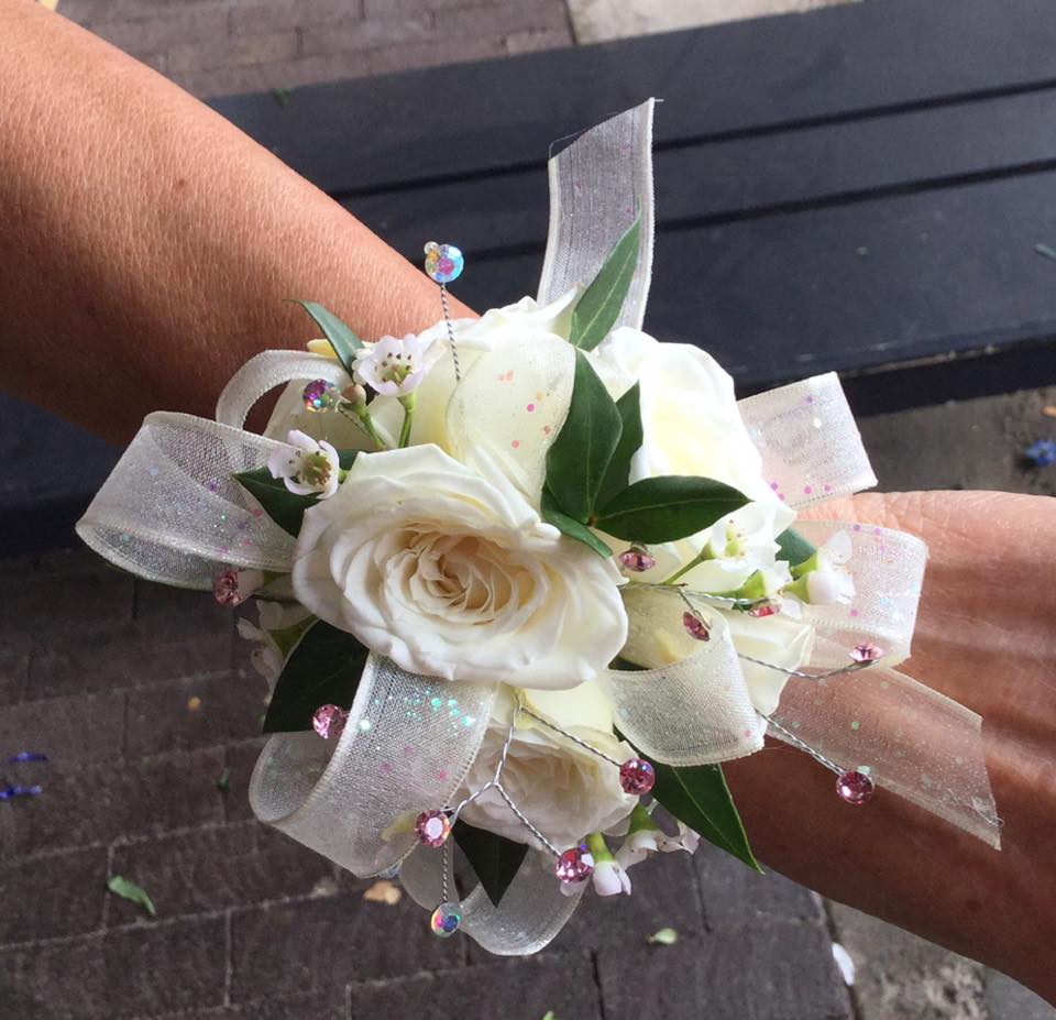 White & Iridescent Wrist Corsage please call store White & Iridescent Wrist Corsage please call store - Our signature custom made wrist corsage with white spray roses and accented with iridescent ribbon and wax flower set on an iridescent Slap wristlet. Also contains add-on iridescent rhinestones We have a wide range of ribbon options and rose colors to choose from. Feel free to visit us at our shop to pick out a custom corsage for you.