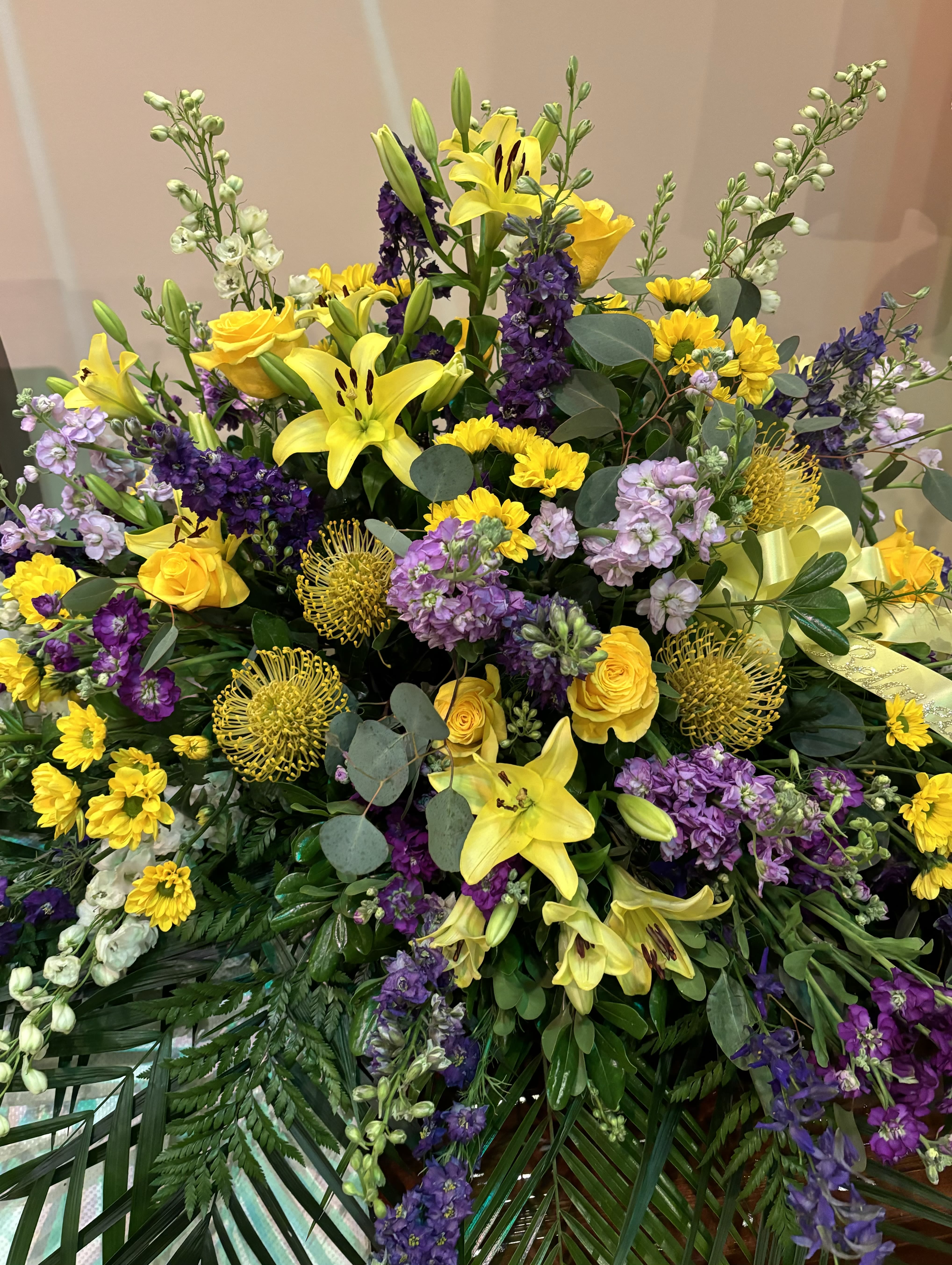 Your light lives on - A bright and beautiful casket spray created with yellow asiatic lilies, protea, roses, purple asters and stock