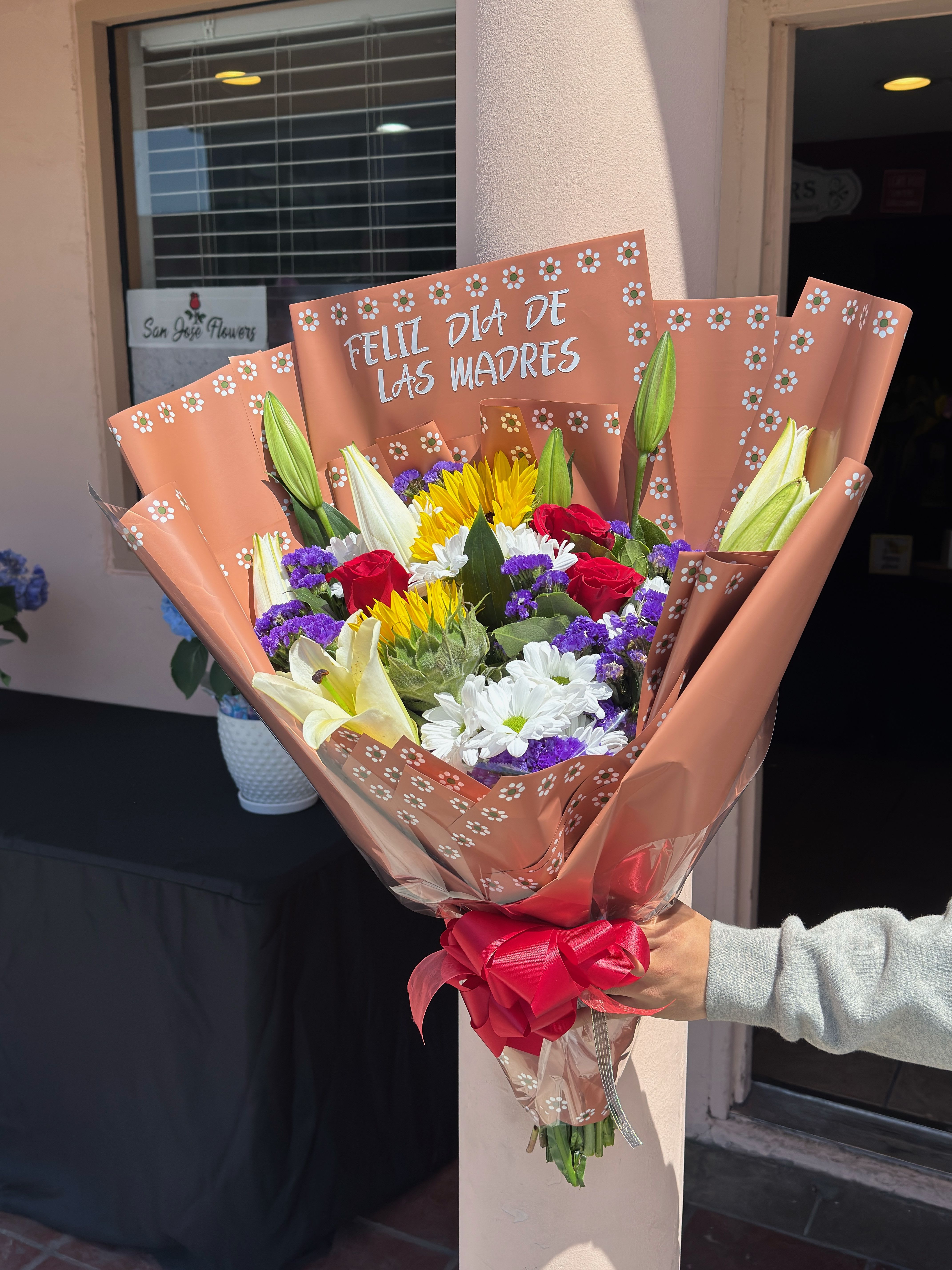 Dia de Mama - Celebrate Mom with this joyful mix of sunflowers, roses, lilies, daisies, and statice — wrapped in barro paper adorned with the words “Feliz Día de las Madres”. Bursting with color and love, it's a bright hug in bouquet form.