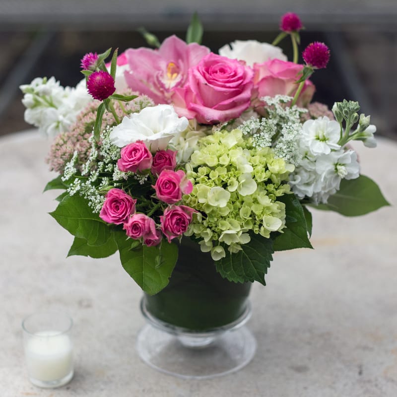 Glass Pedestal Mix - Hydrangeas, spray roses, cymbidium orchids, Queen Ann's lace, lisianthus, lemon leaf, and rosita vendetta roses in a glass pedestal. 
