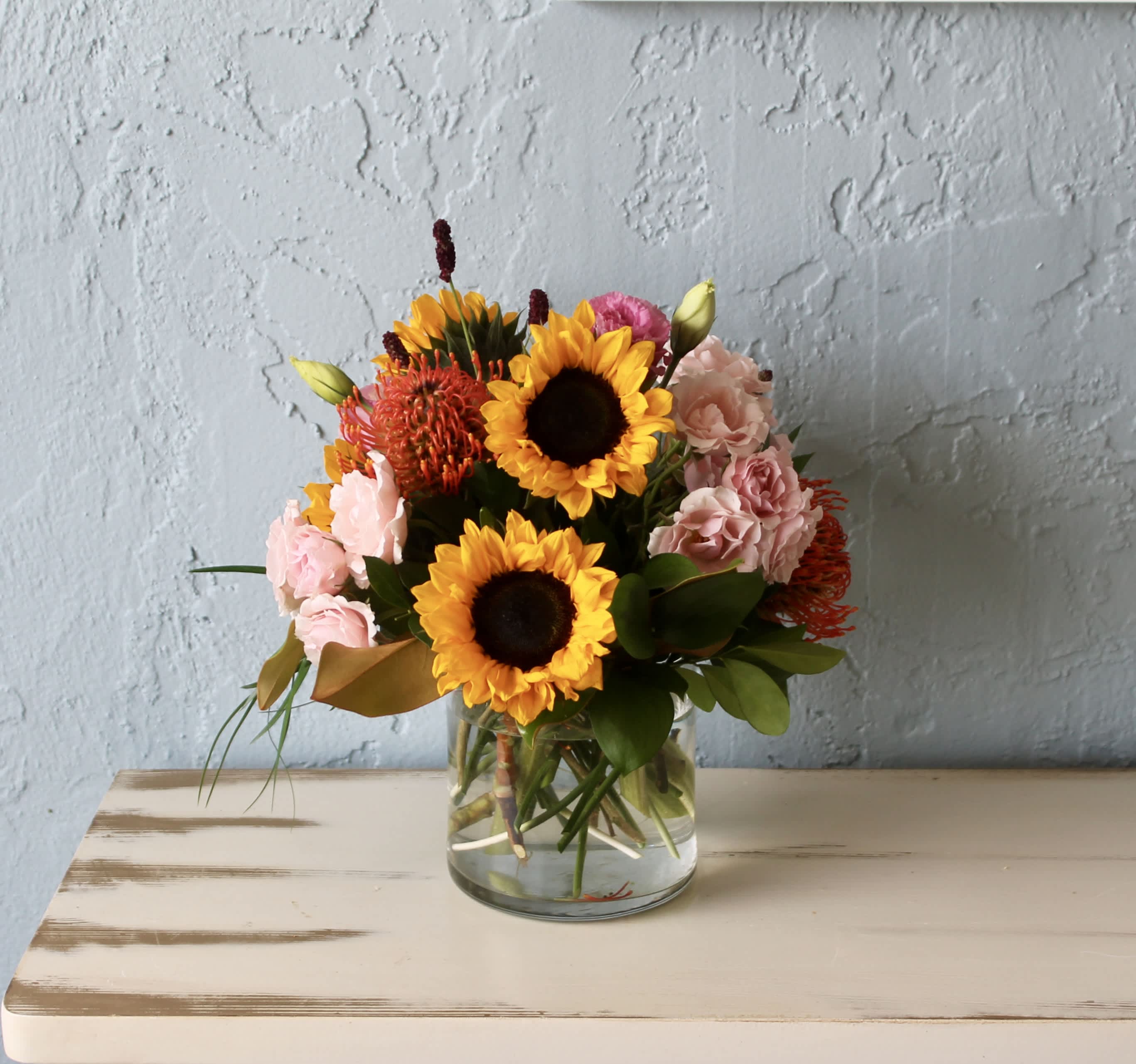 Sunny Day - Summer by the door! Everybody loves a sunshine ! Featuring sunflowers, a pincushion protea, and majolica spray roses, this arrangement is a great option to send to somebody celebrating the summer season.  Approximate Dimensions: 12&quot;D x 8&quot;H