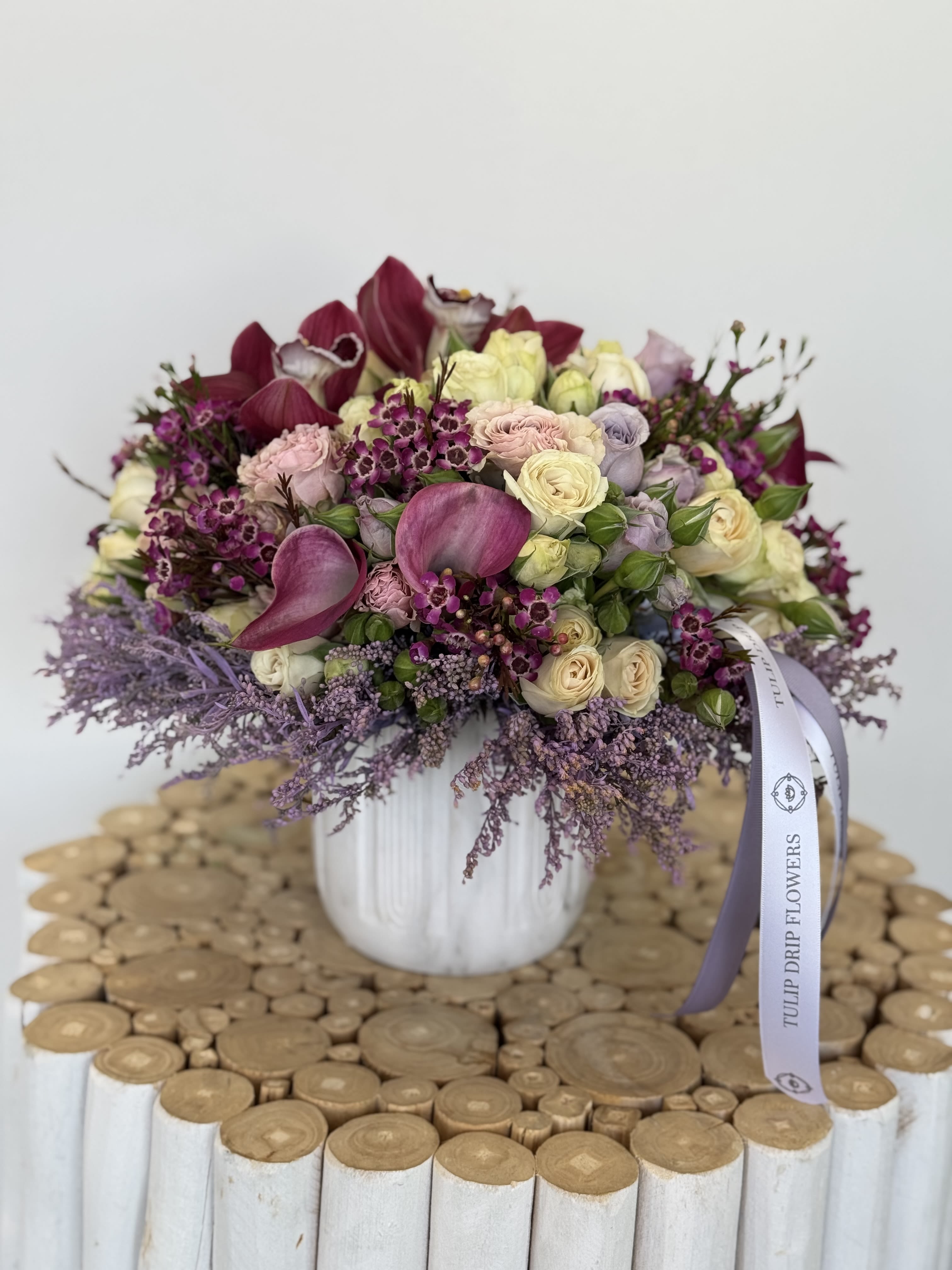 Amethyst Whispers - A stunning vase arrangement featuring calla lilies, orchids, roses, and wax flowers in soft lavender, blush, and cream tones, elegantly tied with a lilac ribbon. Ideal for anniversaries, luxury gifts, birthday surprises, and home décor. The combination of orchids and roses makes this a unique and luxurious floral statement.