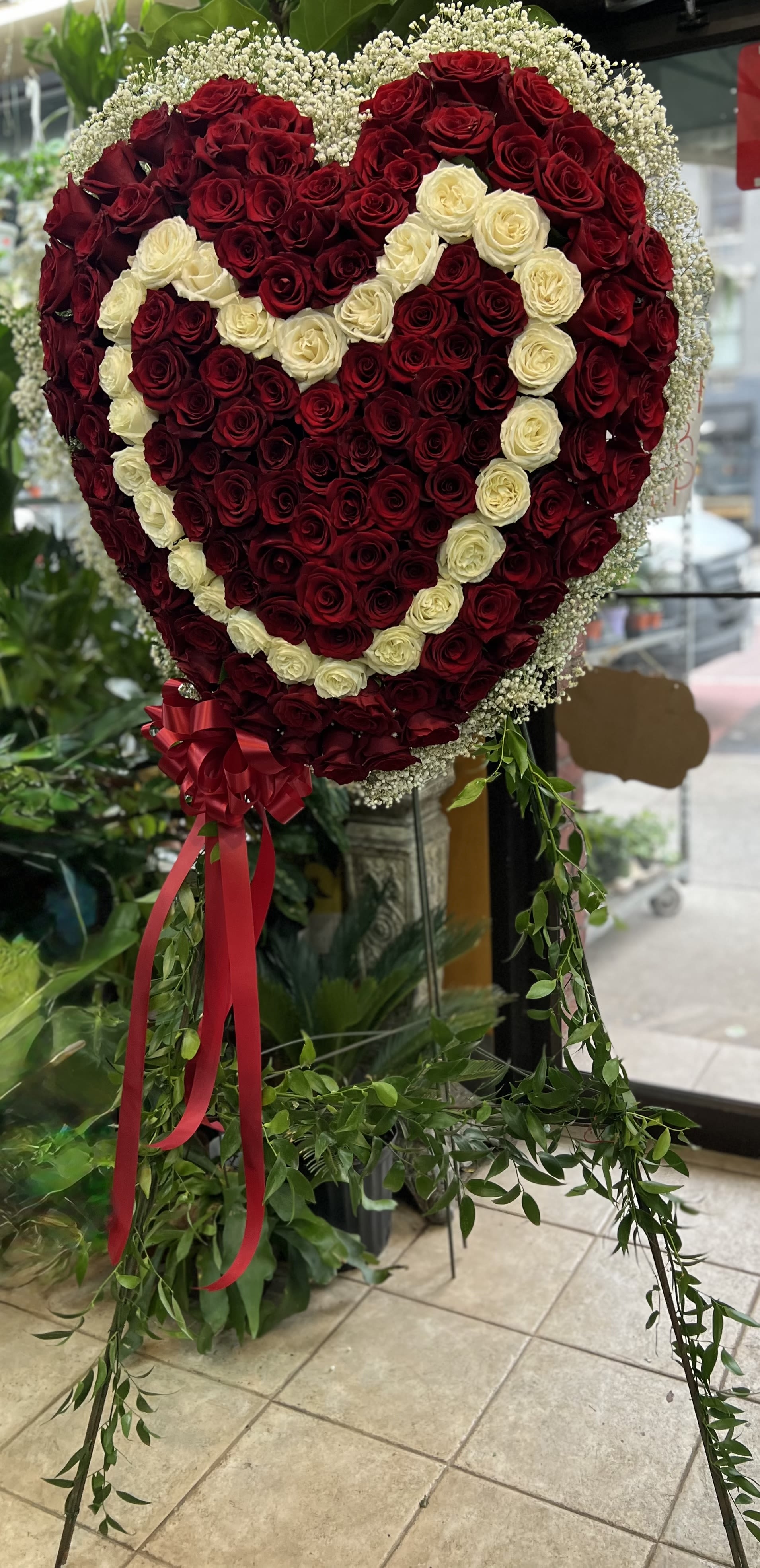 Heartfelt Love Standing Spray Heartfelt Love Standing Spray - A striking and romantic floral tribute, this heart-shaped standing spray is designed to honor love and devotion with timeless elegance. Crafted with lush, velvety red roses as the primary base, the heart is beautifully outlined with pristine ivory roses forming a second heart within — symbolizing love within love. The arrangement is delicately framed with soft baby’s breath, adding a cloud-like softness to the design. Finished with a flowing red satin bow and accented with cascading Italian ruscus greenery, this piece stands tall on an easel, making it a beautiful statement at memorials, anniversaries, or romantic tributes.