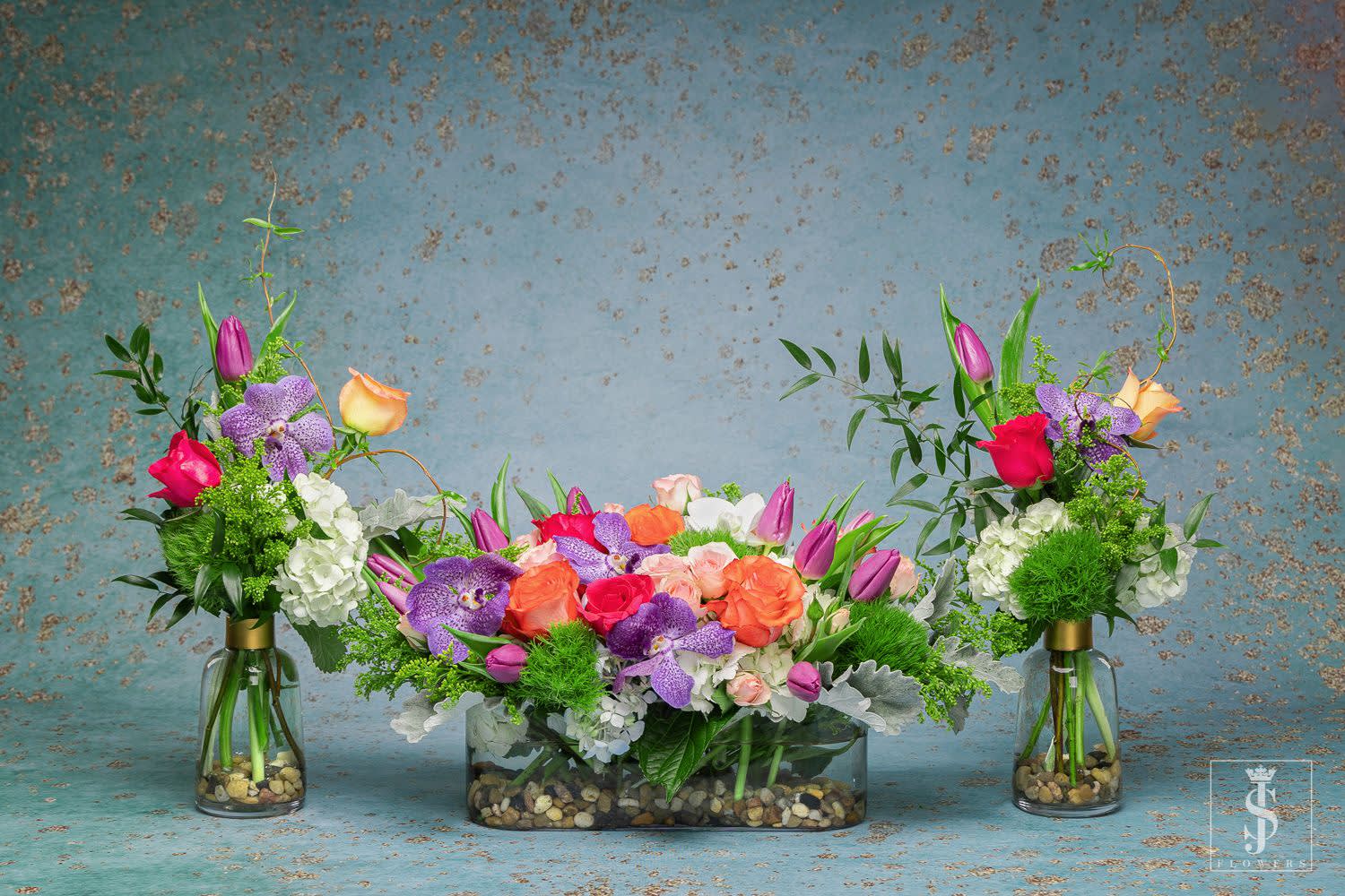 Dinner Centerpiece Trio  - Bring your dinner table to life with this spring trio set centerpiece. This arrangement comes with two bud vases and a long and low clear vase. This set of flowers can all be used on the same dinner table or on other tables in the hosing room. This richly textured design of blue and white hydrangea, lavender hyacinth, peachy, hot pink and light pink roses, tulips, salmon or white ranunculus, and beautiful purple vanda orchis and white orchis . Accented with seeded eucalyptus and dusty miller. 
