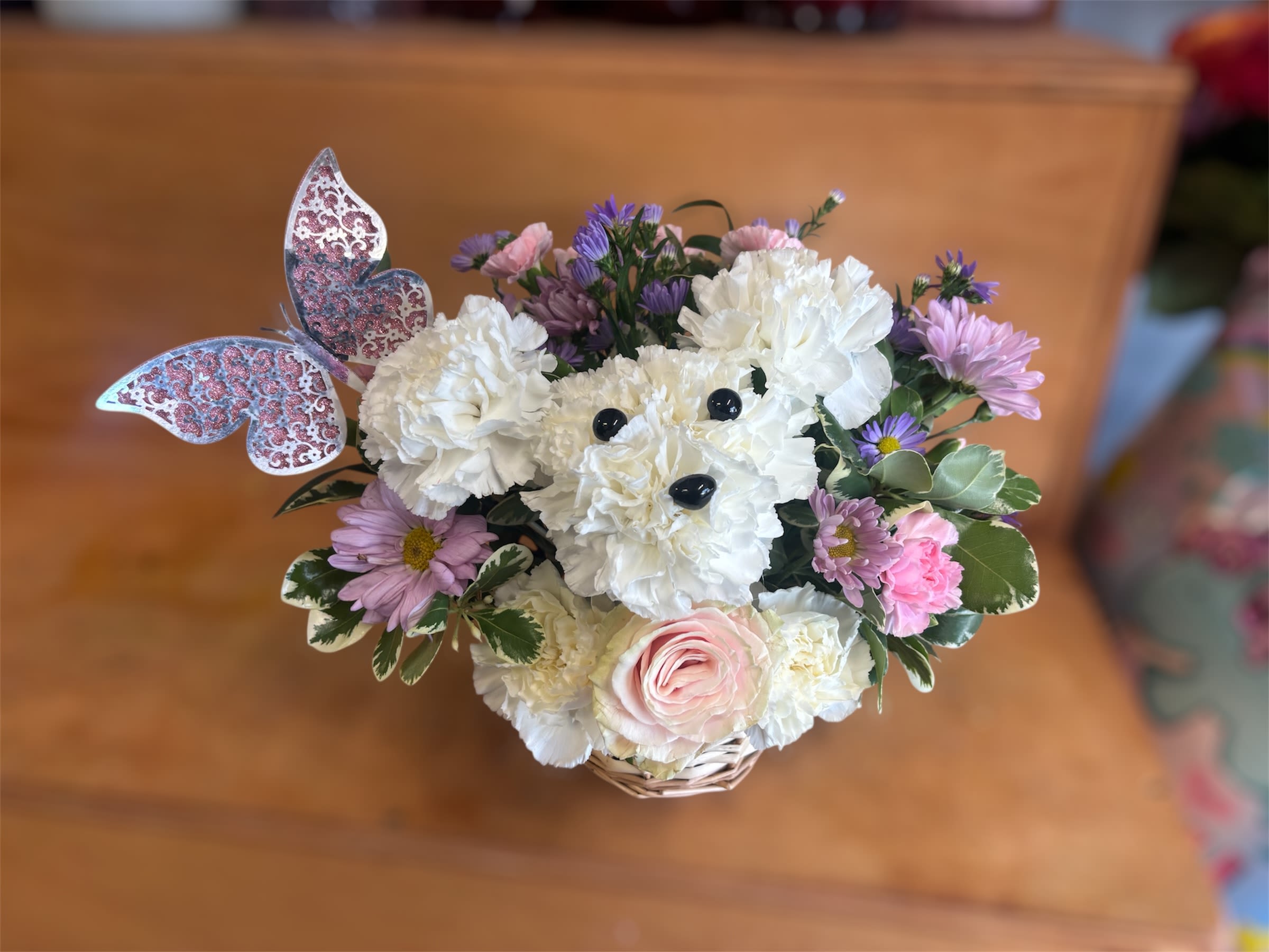 Precious Pup - Our truly original 3D pup is a precious Mother’s Day surprise. Crafted from white carnations, with sweet pink blooms all around and a butterfly accent, this canine creation is designed in a dog bed basket. For more happy wishes, add our festive “Happy Mother’s Day” balloon.  One-sided, 3D arrangement with white carnations crafted in the shape of a dog complete with eyes and nose; surrounded by a mix of pink roses, light pink mini carnations and purple monte casino; accented with assorted greenery and butterfly pick