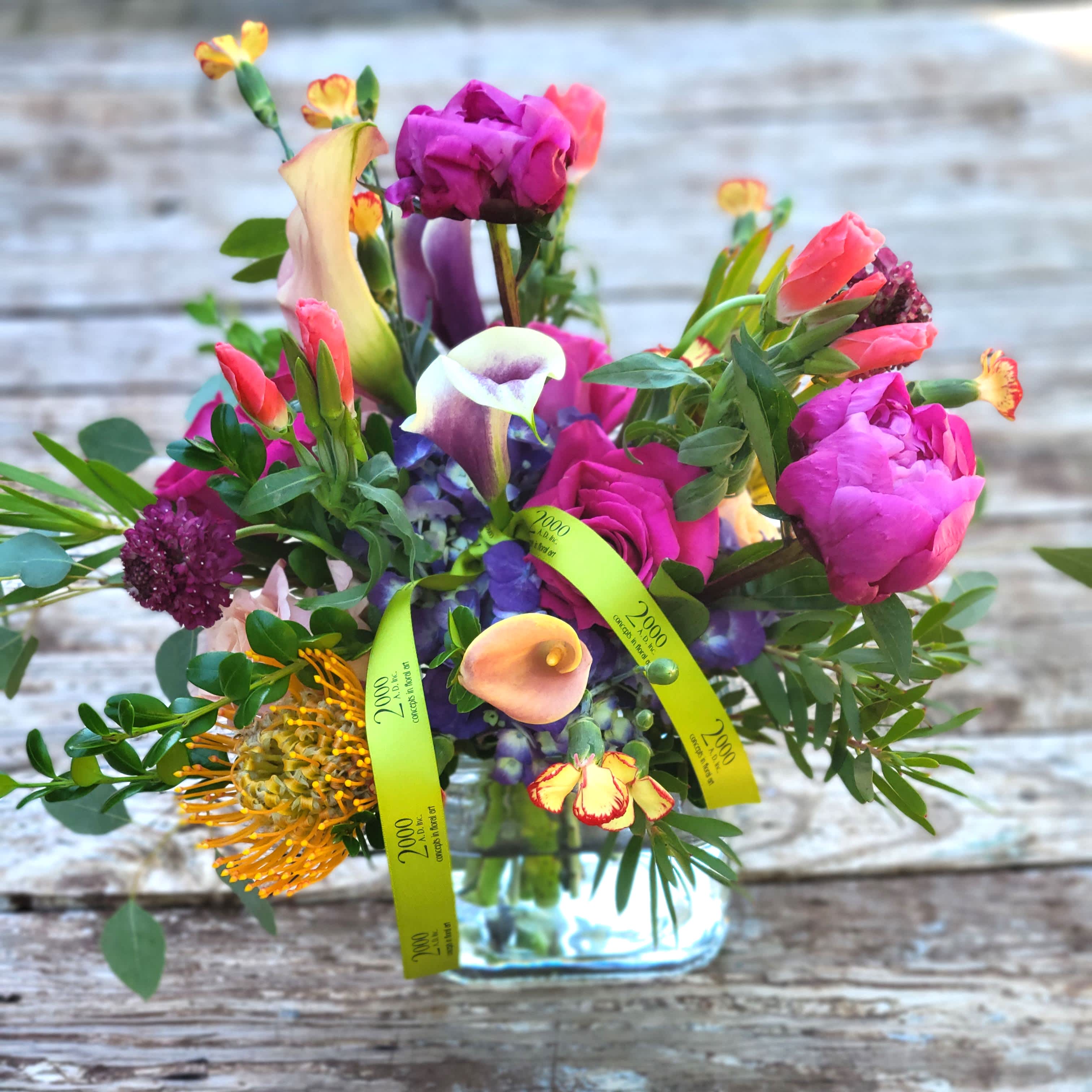 Carnivale - Playful mix of vibrant reds, purples, oranges and hot pink in an energetic arrangement that is sure to please the recipient. Multiple foliages accent the composition. Presented in a clear glass container with exposed stems.