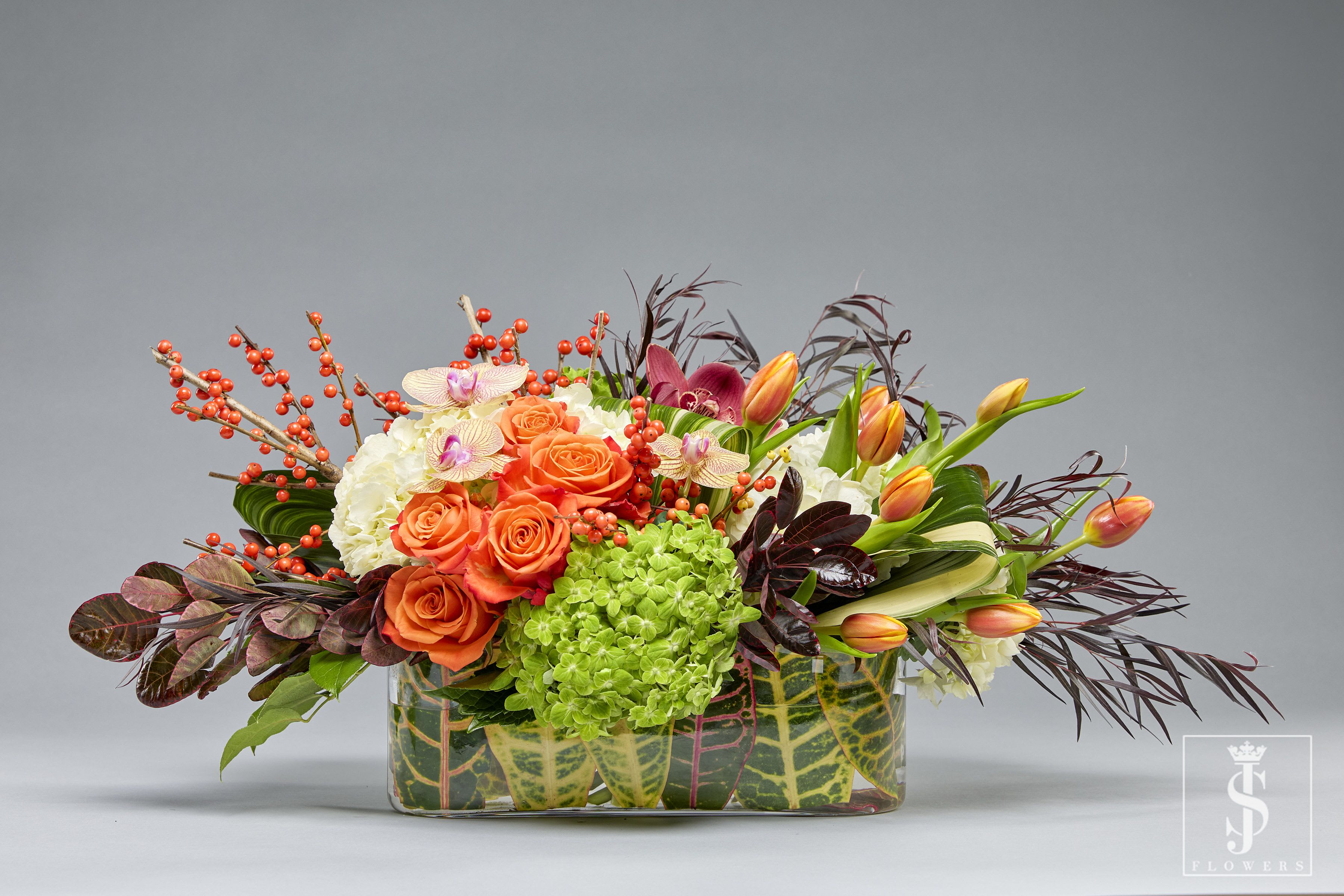 Harvest Bounty Centerpiece - This design is perfect for festive gatherings, Thanksgiving celebrations, or as a thoughtful gift. It captures the essence of the season with its warm, bold hues and natural sophistication.  This exquisite centerpiece radiates warmth and seasonal charm, perfect for celebrating the beauty of autumn. Vibrant orange roses and tulips are the stars of this arrangement, symbolizing joy and vitality. Delicate white hydrangeas add a soft and elegant contrast, while the lush green hydrangeas provide a refreshing touch of vibrancy.  Ornamental foliage in rich burgundy tones adds depth and drama, beautifully complemented by sprigs of hypericum berries in a striking red hue. Accents of phalaenopsis orchids lend a luxurious and exotic flair, creating a captivating blend of textures and colors. The arrangement is thoughtfully presented in a clear vase lined with decorative greenery, adding an organic and earthy feel. 