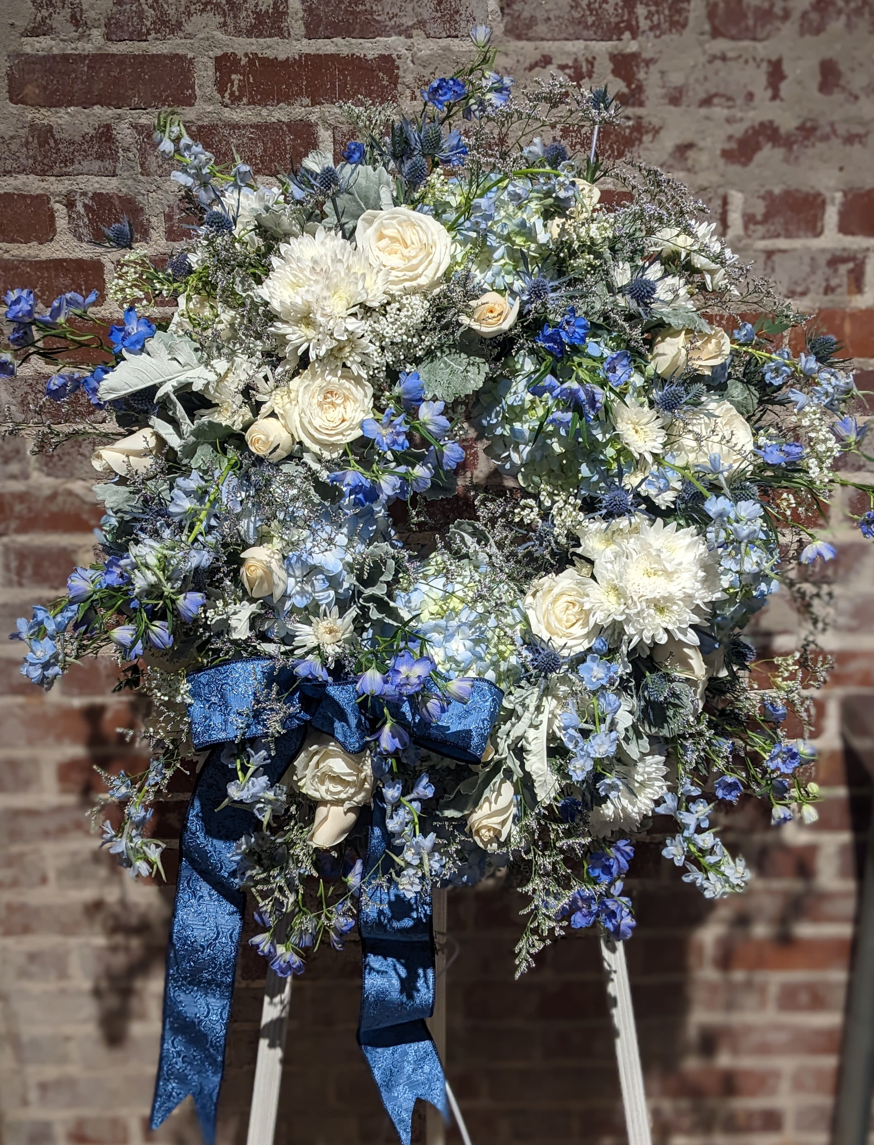 Indigo Cotton Memorial Wreath Indigo Cotton Memorial Wreath - A beautiful floral tribute for the deceased. A lush palette of blue and white blossoms accented with soft green foliages create a sophisticated statement for this celebration of life. Additional color palettes are available upon request. 48 hours notice is required for a color substitution. Please call the shop at 404-314-6194 to discuss.