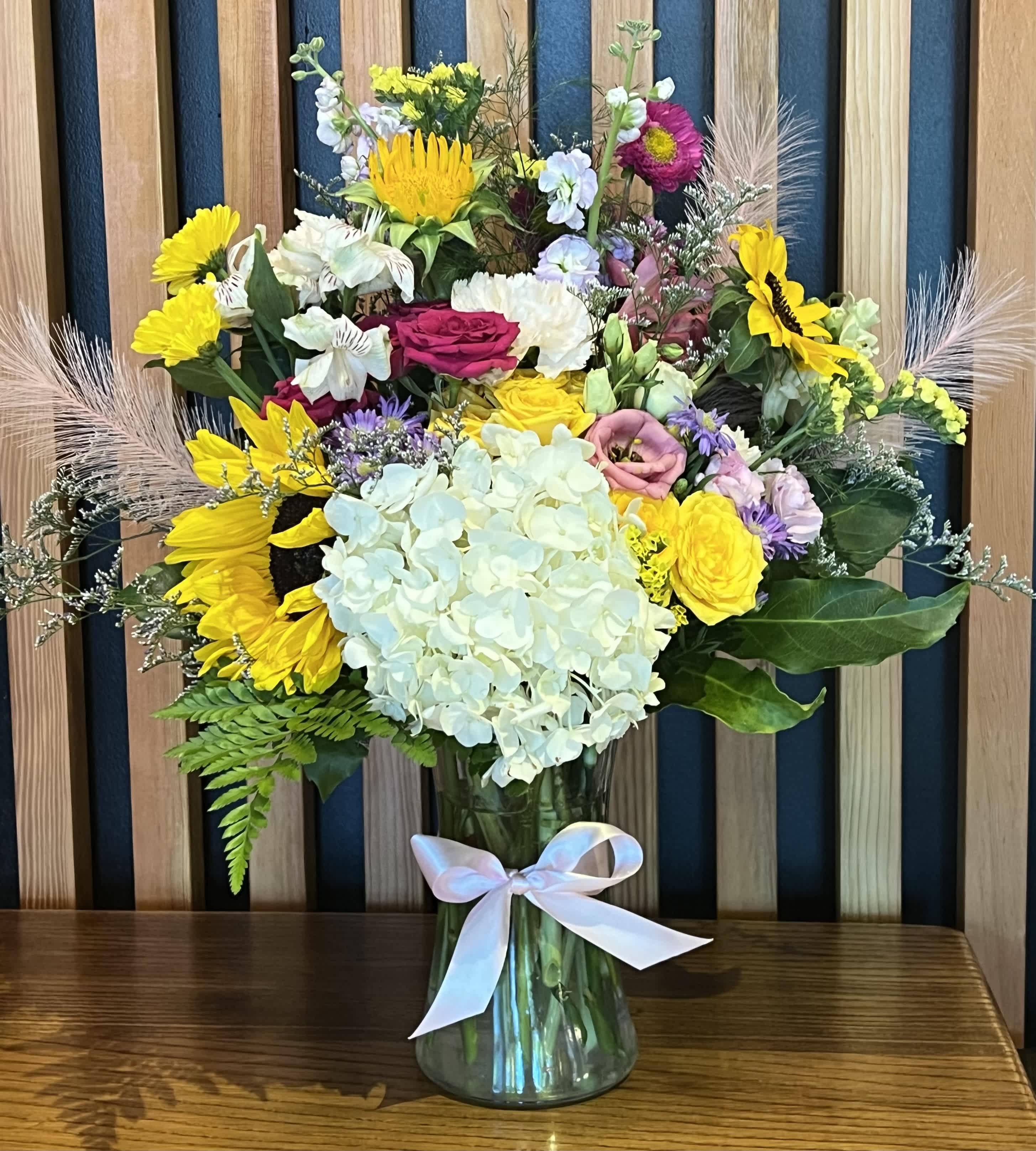 Sun-kissed Garden - A vibrant and cheerful bouquet in a vase featuring sunflowers, spray roses, alstroemeria, stock, and seasonal blooms. The sunflowers, with their large, golden-yellow petals, act as the centrepiece, radiating warmth and positivity.   The bouquet is tied together with lush greenery, framing the flowers and enhancing the overall lush and abundant feel. The result is a harmonious blend of colors, shapes, and textures that capture the spirit of a sun-drenched summer garden.