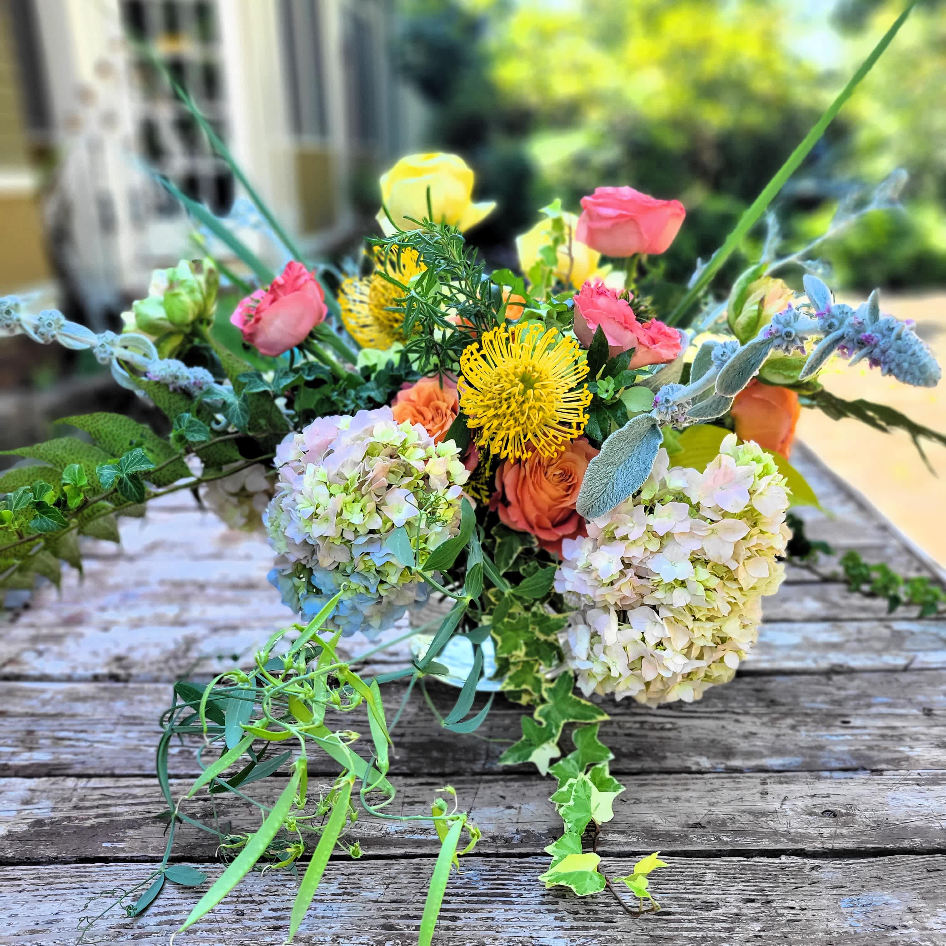 Rebecca - A mixed hydrangea cluster provides the base for this lush arrangements. Unique garden foliage completes the composition which is designed in a clear glass container. Stems are exposed.