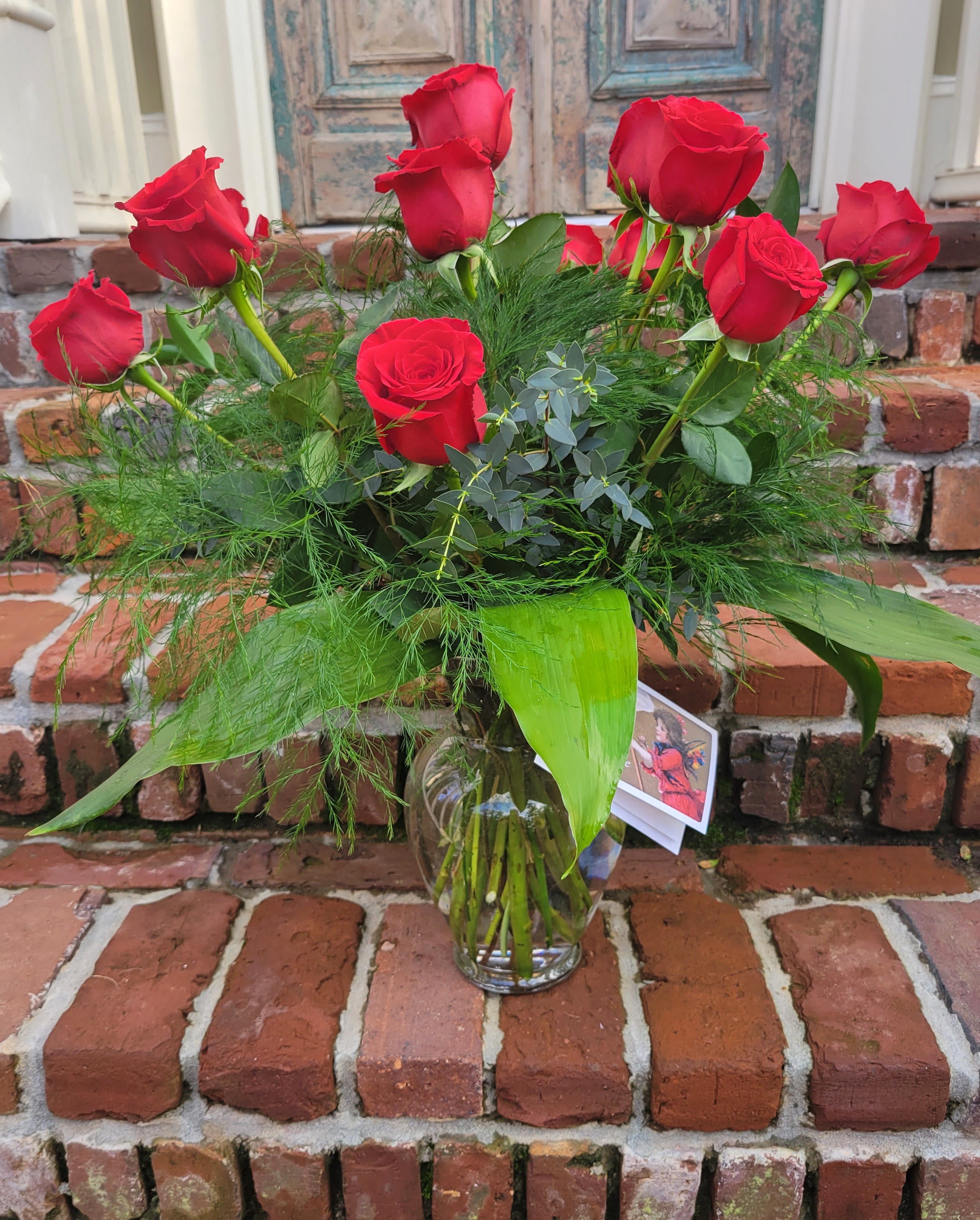 Red Butler - Classic Southern composition of red Freedom roses and lush assorted foliages. Executed in a clear glass container with exposed stems. A vintage Valentine card is included to express your message of love. (Card design will vary.)