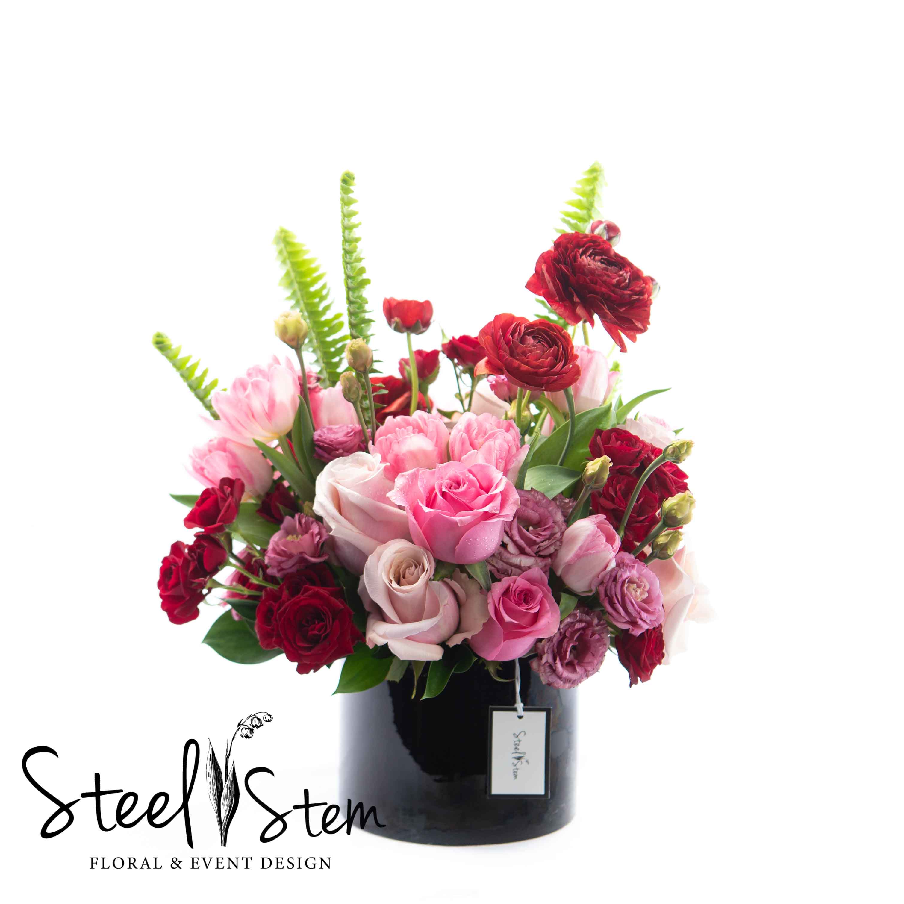 Blooming Love Blooming Love - A classic black ceramic cylinder is designed using shades of pinks and deep burgundy flowers for a warm and feminine feel. Shades of roses, elegant ranunculus, fluttering with tulips, and so much more are used in this abundant Mother's Day selection.