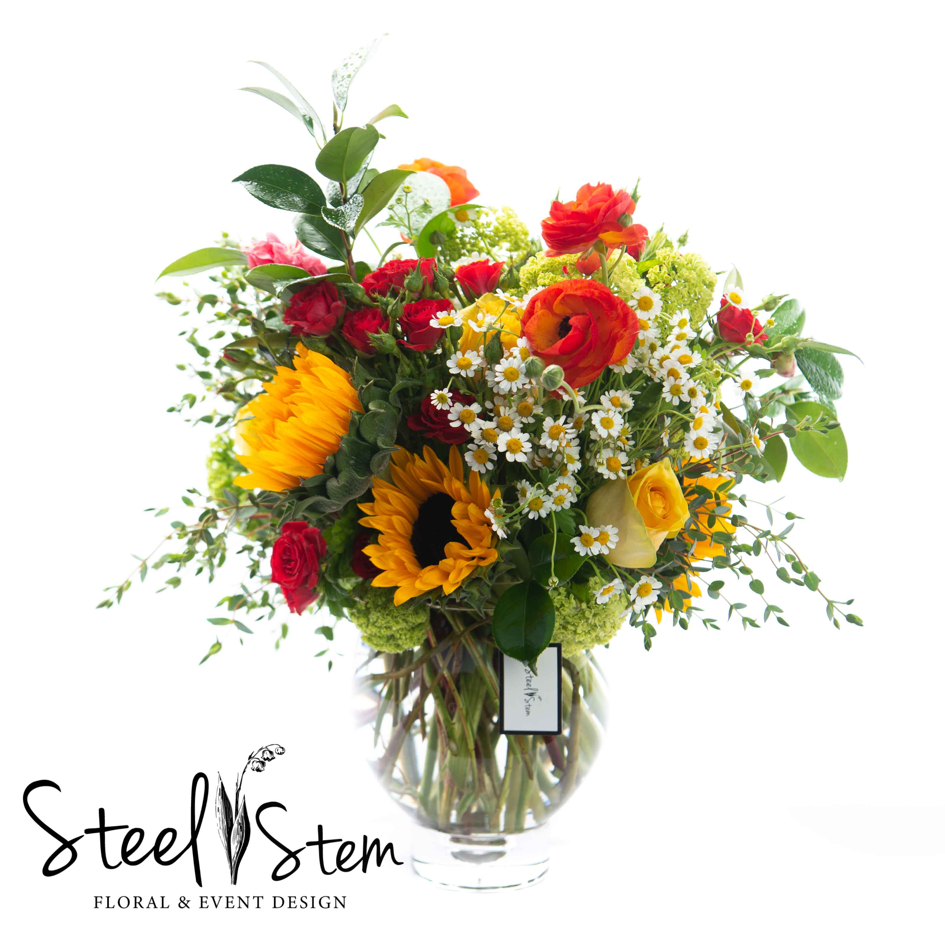 Sun-Kissed Spring  - Vibrant and organic, our Abundance of Spring is an incredibly cheerful and happy arrangement that will lift anyone's spirits! Our lush and full design uses bright yellow sunflowers and roses, notes of chamomile, seasonal orange ranunculus, red spray roses, and a variety of our favorite garden greeneries that rests inside of a glass fish bowl.   Measures approx. 12&quot; W x 20&quot;H