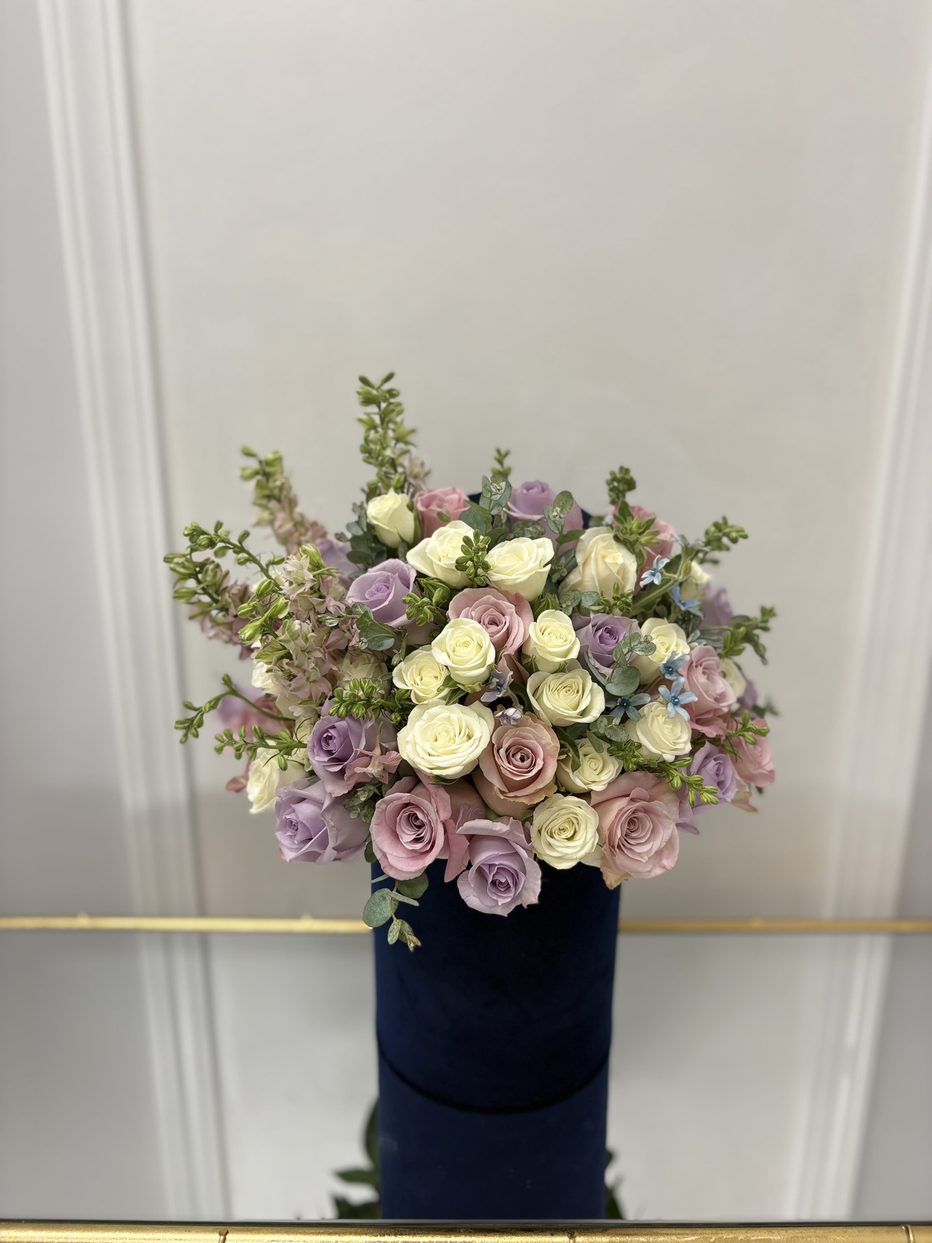 Miss Pastel In Blue Hat Box BY VLM Flower - Light pink and lavender roses with larkspur and white spray roses.