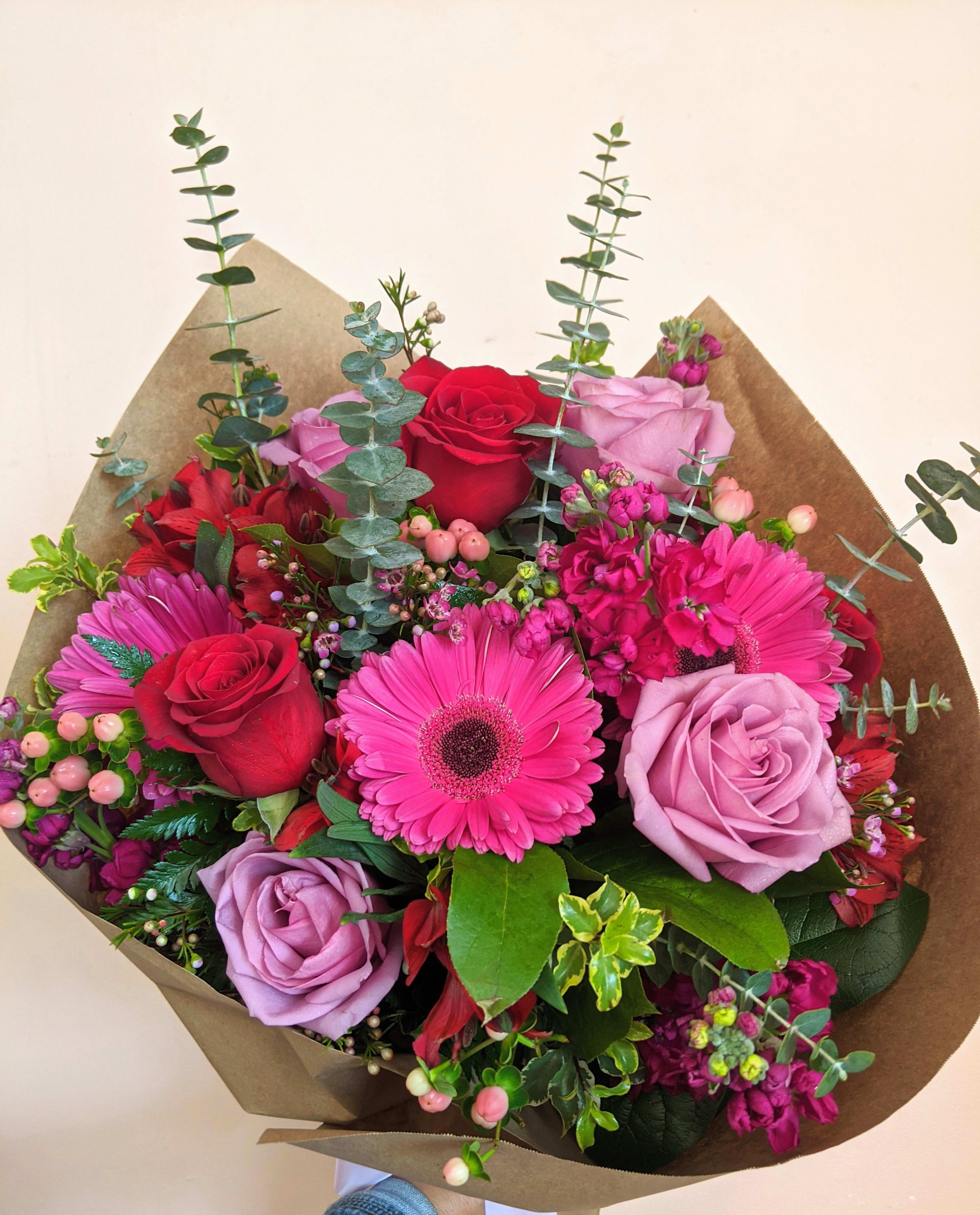 Secret Garden Wrapped Bouquet - Our talented designer will choose the perfect tones of lavender, fuchsia, and red blooms of roses, gerberas (may be substituted for valentines week with lilies), and alstroemerias nestled in eucalyptus greens plus the perfect filler flower to give this rustic wrapped bouquet the perfect look. 