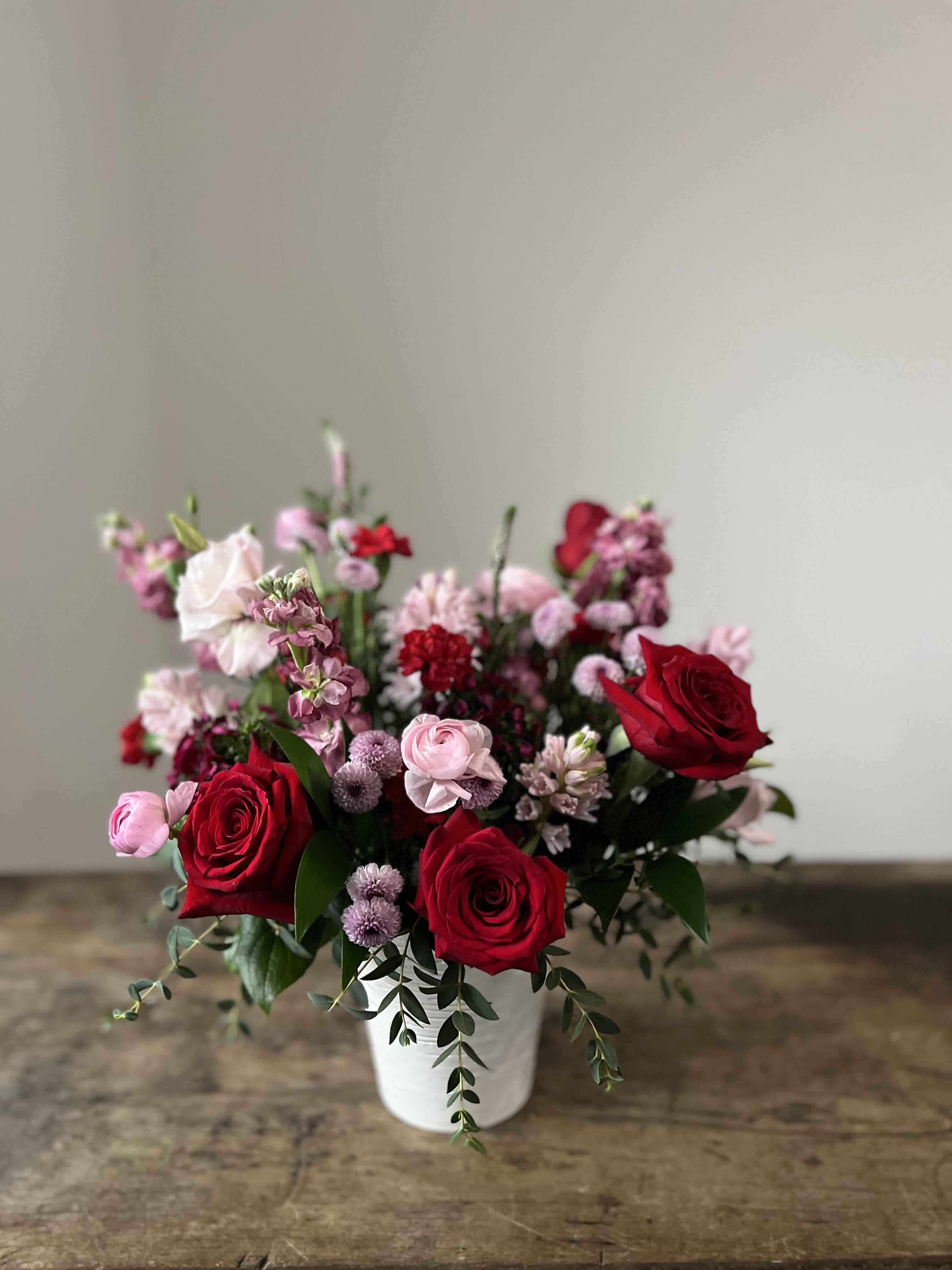 Harmony Vase Arrangement Harmony Vase Arrangement - Elevate any space with this elegant floral design, featuring a harmonious blend of rich red, soft lavender, and deep purple blooms. This sophisticated palette exudes timeless beauty with a modern twist, making it perfect for any occasion. Artfully arranged in a sleek white ceramic pot, the design highlights the contrast between bold colors and minimalist charm. Lush roses, delicate lisianthus, and striking stock are complemented by hyacinth, ranunculus, and textural greenery, creating a balanced and refined composition. Approximately 9” x 11”