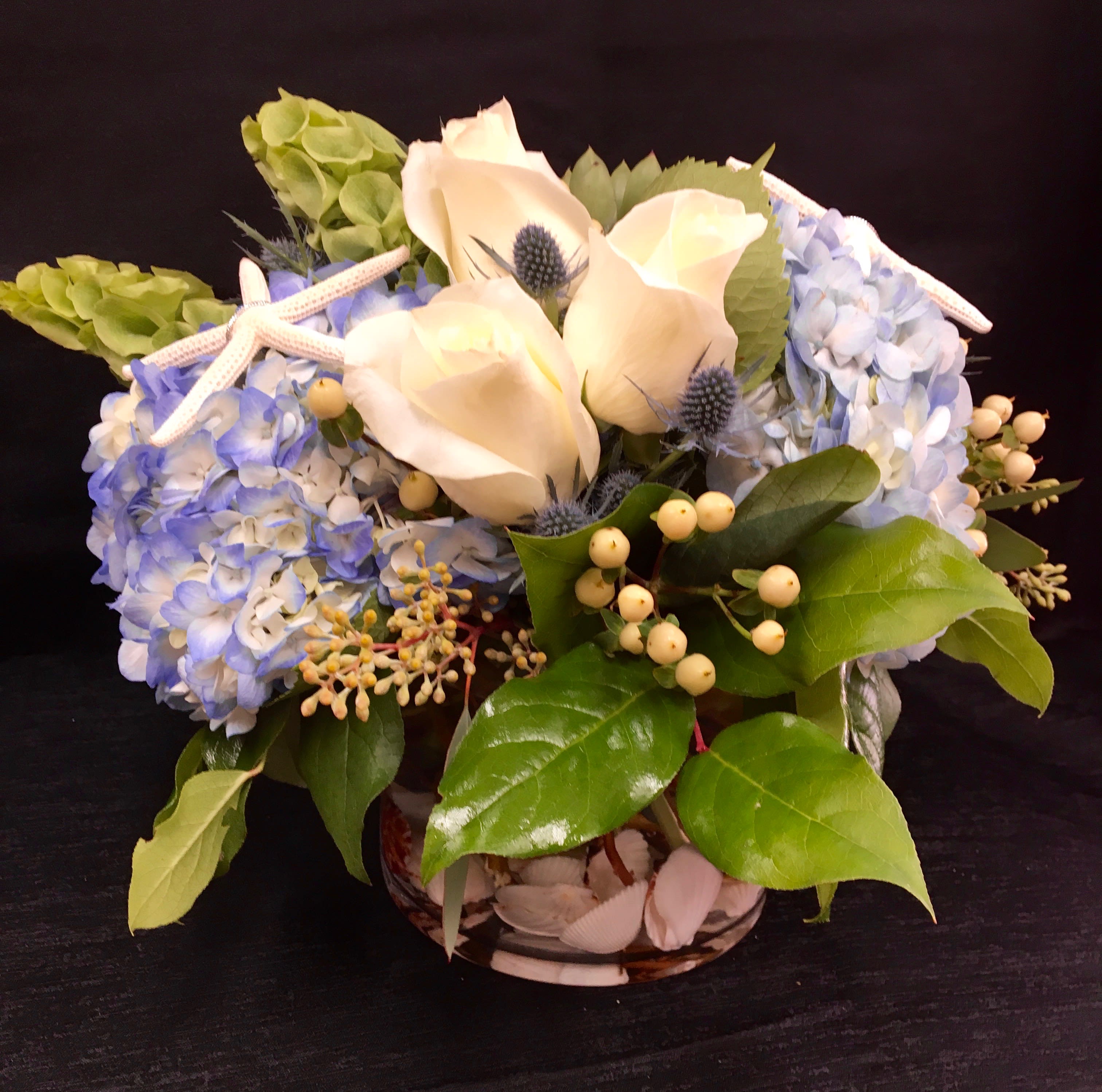 BLUE BEACH PARADISE - This beautiful arrangement has roses, hydrangea, thistle, star shells, cream coffee beans, bells of Ireland in a glass bubble bowl with shells. The perfect arrangement for La Jolla.
