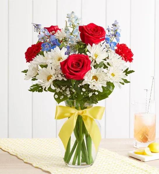 Tie a Yellow Ribbon - All-around arrangement with red roses and carnations, white daisy poms, and blue delphiniums, accented with baby’s breath and assorted greenery Artistically designed in a clear glass vase accented with a yellow satin ribbon;