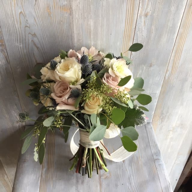 Happily Ever After - Indulge in the romantic beauty of a bridal bouquet that features delicate mauve and pristine white roses, accentuated by the textured charm of seeded eucalyptus and the striking pop of blue thistle. Walk down the aisle with grace and sophistication with this stunning arrangement. 