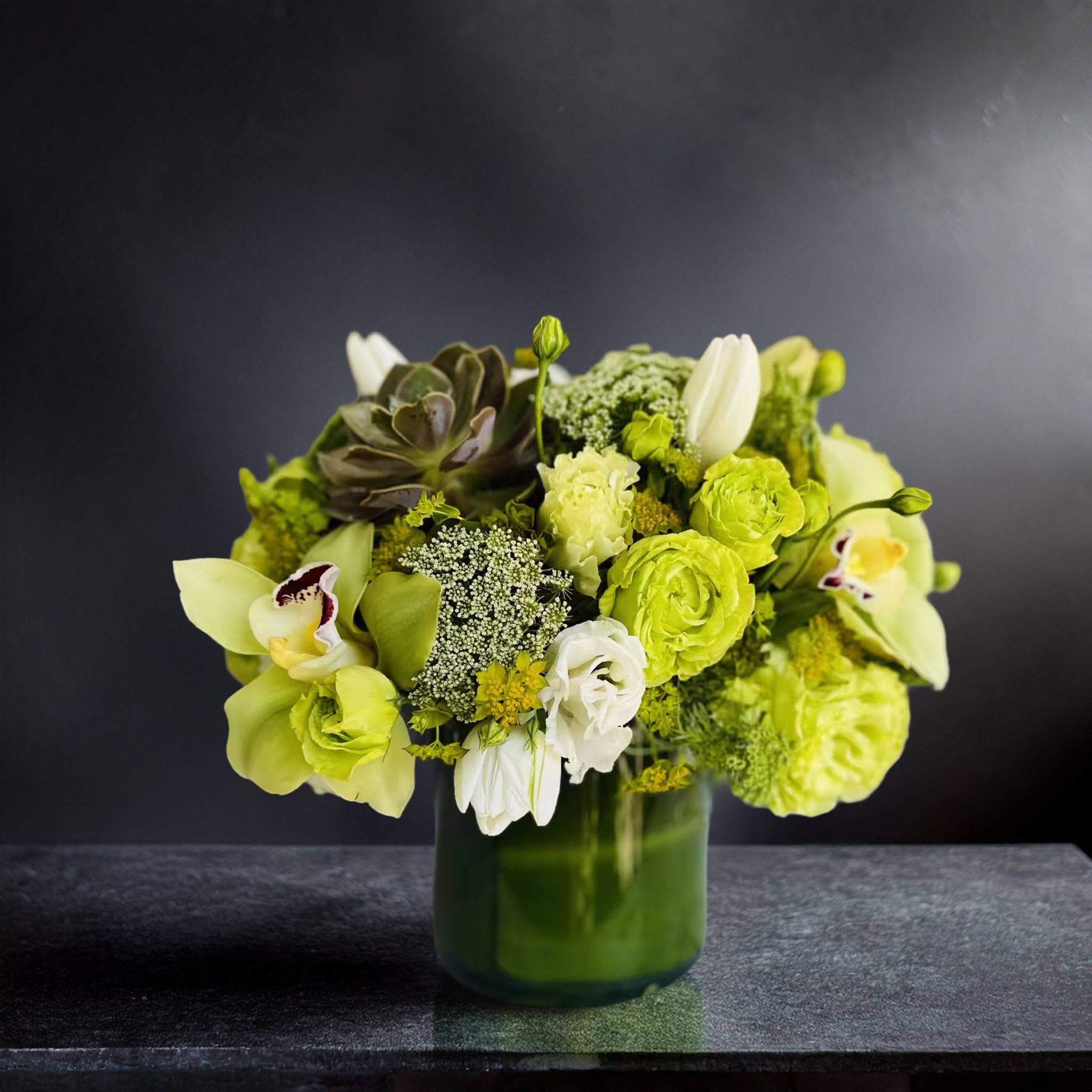 Garden Of Green <3 Garden Of Green <3 - Bold and contemporary with a clean botanical elegance, this green and white floral arrangement commands attention with its tonal harmony and refined composition. Set in a glossy dark green cylinder glass vase, this design features vibrant chartreuse roses, lime green hydrangeas, green cymbidium orchids with burgundy throats, creamy white tulips, and ruffled lisianthus. The centerpiece is accented with Queen Anne’s lace and a structured succulent, adding texture and modern flair. The moody charcoal backdrop enhances the richness of the greens and whites, making this design feel both luxurious and artful — perfect for high-end environments and design-savvy clients. This arrangement is ideal for luxury home staging, corporate gifting, hotel suites, or architectural spaces that lean into minimalist or monochrome aesthetics. It’s also a favorite for those who appreciate modern floral design with an organic edge. Whether delivered as a gesture of gratitude or placed as a signature decor piece, it delivers sophistication with a fresh, serene palette. Touch of Orchids LA offers same-day delivery of this green and white orchid-focused arrangement throughout Los Angeles, including Beverly Hills, Brentwood, West Hollywood, Culver City, and Century City. A staple of our modern collection, this arrangement elevates any interior with ease.
