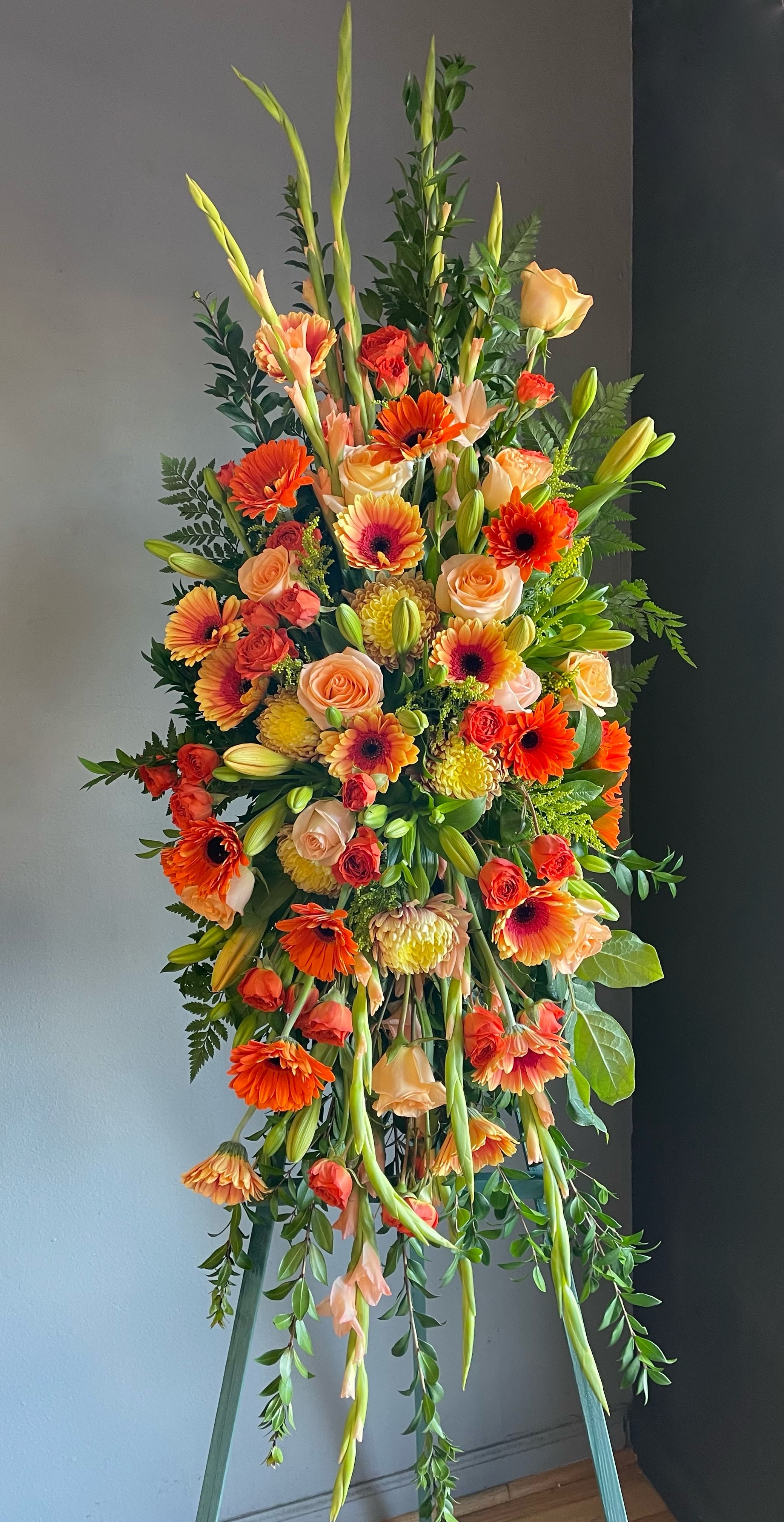 FUNERAL STANDING SPRAY - Peach Dreams 81 - This is a beautiful option for a funeral service, church or graveside. This standing spray is a one sided flower arrangement on a 5’ easel (easel included) displaying a spray approximately 4’ tall and 2’5” wide. This lovely mix of peach and orange flowers of gerber daisies, roses, gladiolas, and lilies is a great option to remember that sunny person in your life.   **This is a piece for a funeral or memorial service. Please place the order at least 2 days prior to the service to guarantee a certain arrangement. Anything sooner and we cannot guarantee it will match the picture. These pieces can be customized to the color of your choice. The flowers may vary depending on availability of the flowers, special requests and quality of the flowers we have in stock. If you have a special request please add it to our special instructions when placing your order. If you have any questions please don’t hesitate to call. 714 289 1123**