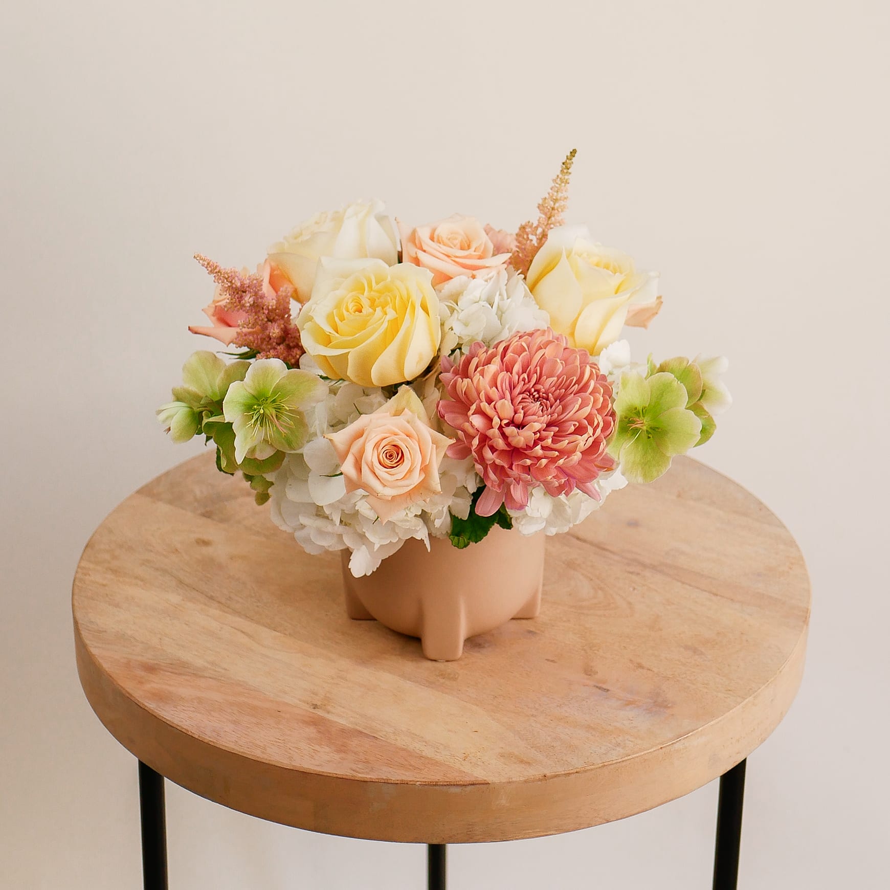 Lacey - This flower arrangement showcases a soft and warm palette of ivory and peach tones. Featuring our premium Candlelight roses, peach baby roses, hellebores, blush astilbe and dusty-rose chrysanthemums, it blends delicate textures in a modern beige ceramic container.  This warm and inviting arrangement is perfect for Mother's Day, symbolizing love, gratitude, and admiration. Despite its compact design, it’s filled with premium blooms like the beloved Candlelight rose—our largest rose, cherished for its soft fragrance and elegant beauty. Complimented with lush Linette mums known for their impressive longevity, this arrangement offers a lasting impression of grace and affection.