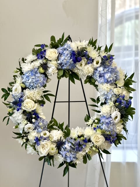 Ocean Breeze Sympathy Wreath by Westford Florist Art by Veny - The Ocean Breeze Sympathy Wreath by Westford Florist Art by Veny is a stunning tribute designed to convey heartfelt sentiments during times of loss. This 18-inch open oasis wreath features a serene palette of delicate blue hydrangea, striking blue delphinium, and vibrant blue thistle, harmoniously blended with elegant white roses, white lisianthus, poms, and carnations. Accents of Italian ruscus and variegated pitt greens enhance its natural beauty, creating a soothing presence that offers comfort and remembrance. Perfect for memorial services or as an expression of sympathy sent directly to the home, this exquisite arrangement serves as a poignant reminder of love and cherished memories in challenging times.