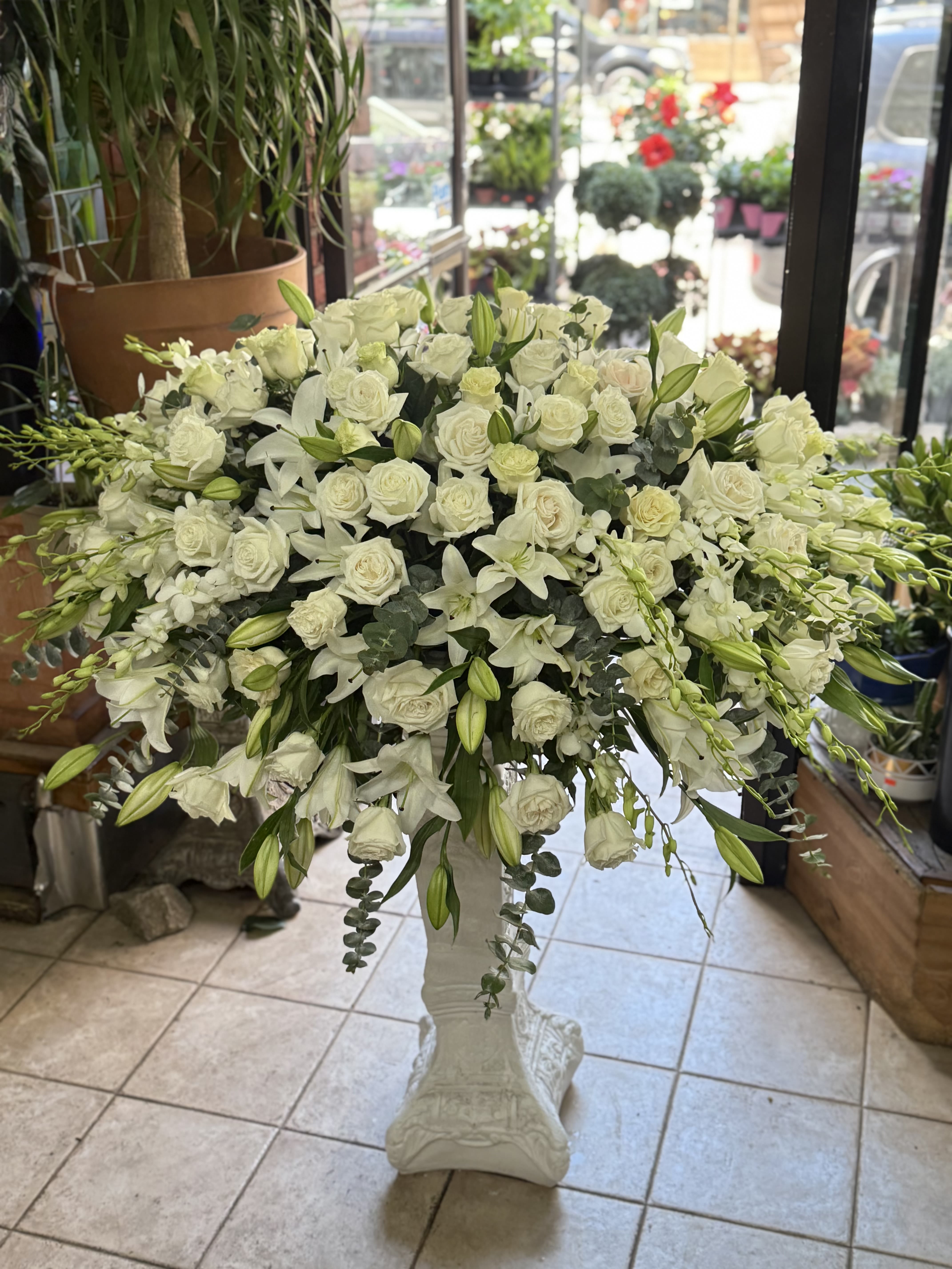 Eternal Elegance  - An elegant expression of love and remembrance  Honor a cherished life with the timeless beauty of our Eternal Elegance Casket Spray, a stunning arrangement of pure white roses, lilies, dendrobium orchids, and lush greenery. Designed with elegance and reverence, this classic spray rests gracefully atop the casket, offering a serene tribute that symbolizes peace, purity, and eternal love.  Ideal for honoring a loved one with a dignified and heartfelt floral expression, this arrangement is handcrafted with the utmost care and attention to detail.  Please Note: The decorative pedestal stand shown in the image is not included and is for display purposes only.