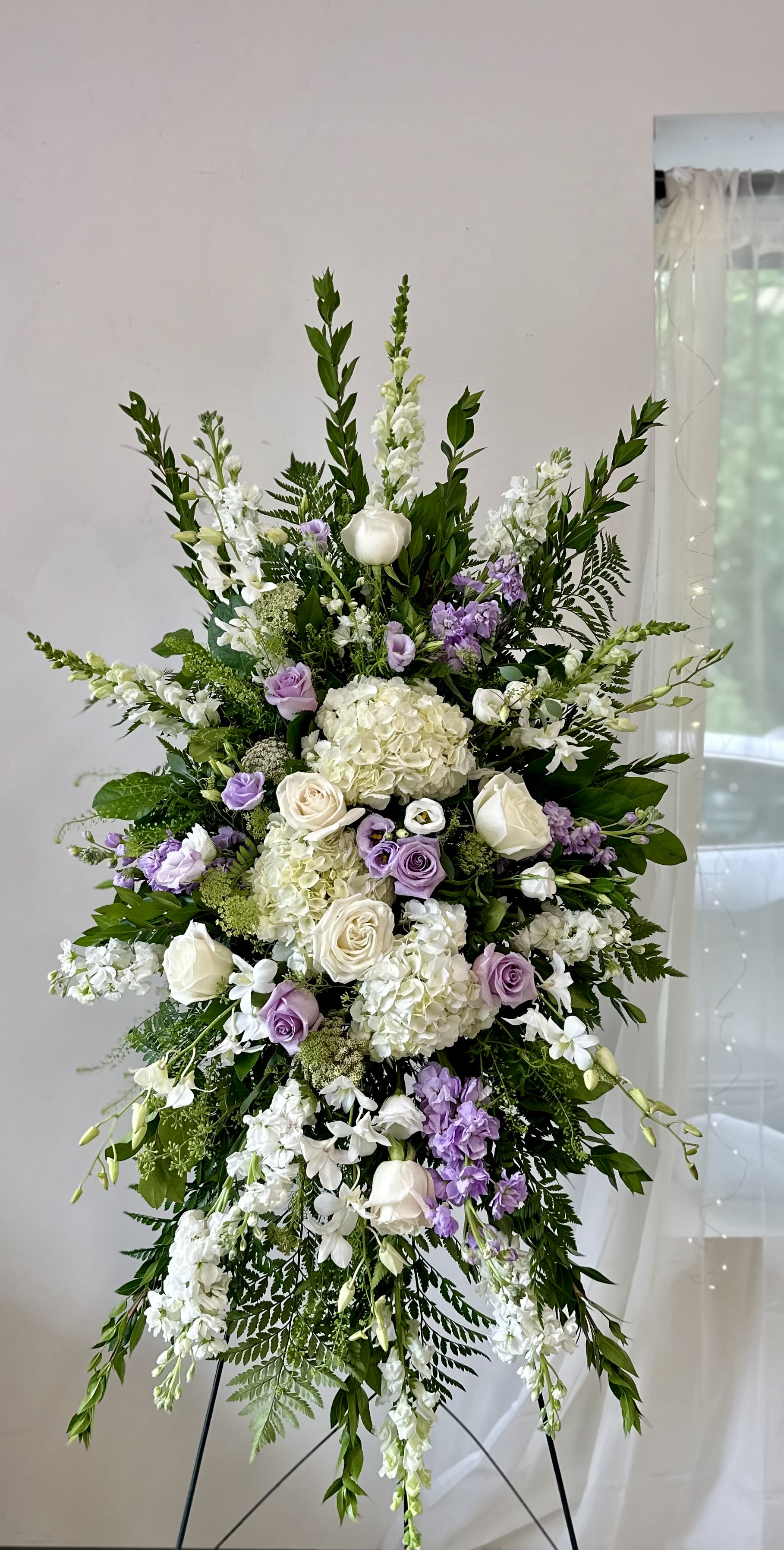 Lavender Serenity Standing Spray by Westford Florist Art by Veny - Capture the essence of tranquility and compassion with our Lavender Serenity Sympathy Standing Spray by Westford Florist Art by Veny. Thoughtfully arranged, this exquisite display features an elegant high-end blend of pristine white hydrangeas, stock, snapdragons, and roses, harmoniously complemented by soft lavender hues from stock, lisianthus, and delicate orchids. Accentuated with Queen Anne' lace for added grace, this standing spray is a heartfelt tribute designed to convey your deepest sympathies during times of loss. A perfect expression of love and remembrance that brings comfort to those who grieve. Embrace serenity in every petal; let your sentiments bloom beautifully through this stunning floral arrangement by Westford Florist Art by Veny. Picture shows deluxe edition.