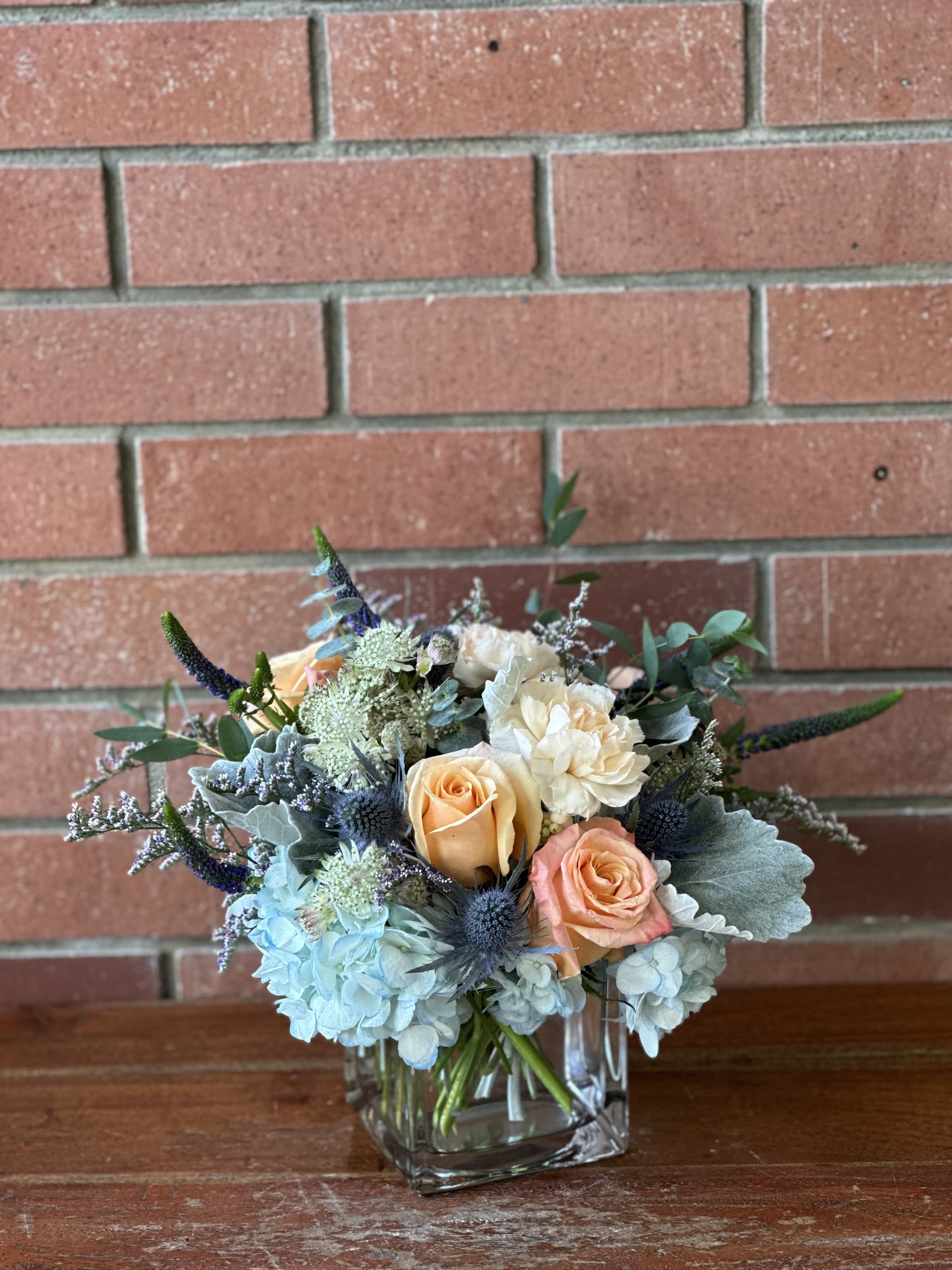 Blue &amp; Peach Modern Cube Bouquet  - Modern clear cube vase filled with blue hydrangea, blue and gray lush greenery eucalyptus and dusty miller, blue thistle, peach roses and carnations, blue filler flowers and astranthia flowers. 