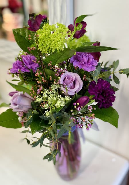 Lavender Lush Lavender Lush - A beautiful blend of vibrant purple blooms arranged with green accents and mixed greenery into a tall, narrow clear glass vase.