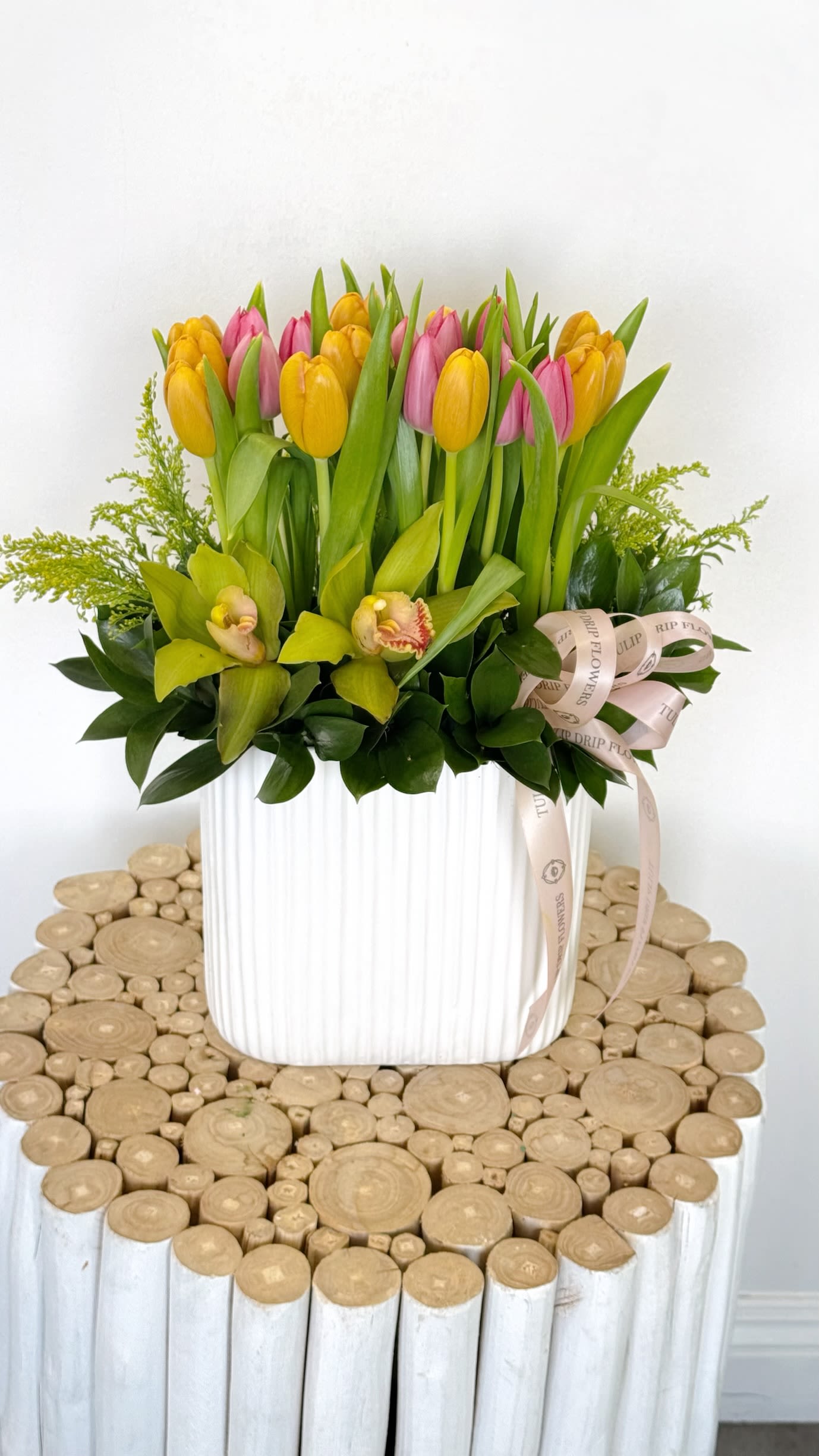 Spring Tulip Garden Spring Tulip Garden - A cheerful design featuring yellow and pink tulips with green cymbidium orchids and lush greenery in a tall white vase. A bright and fresh Mother’s Day flower gift.