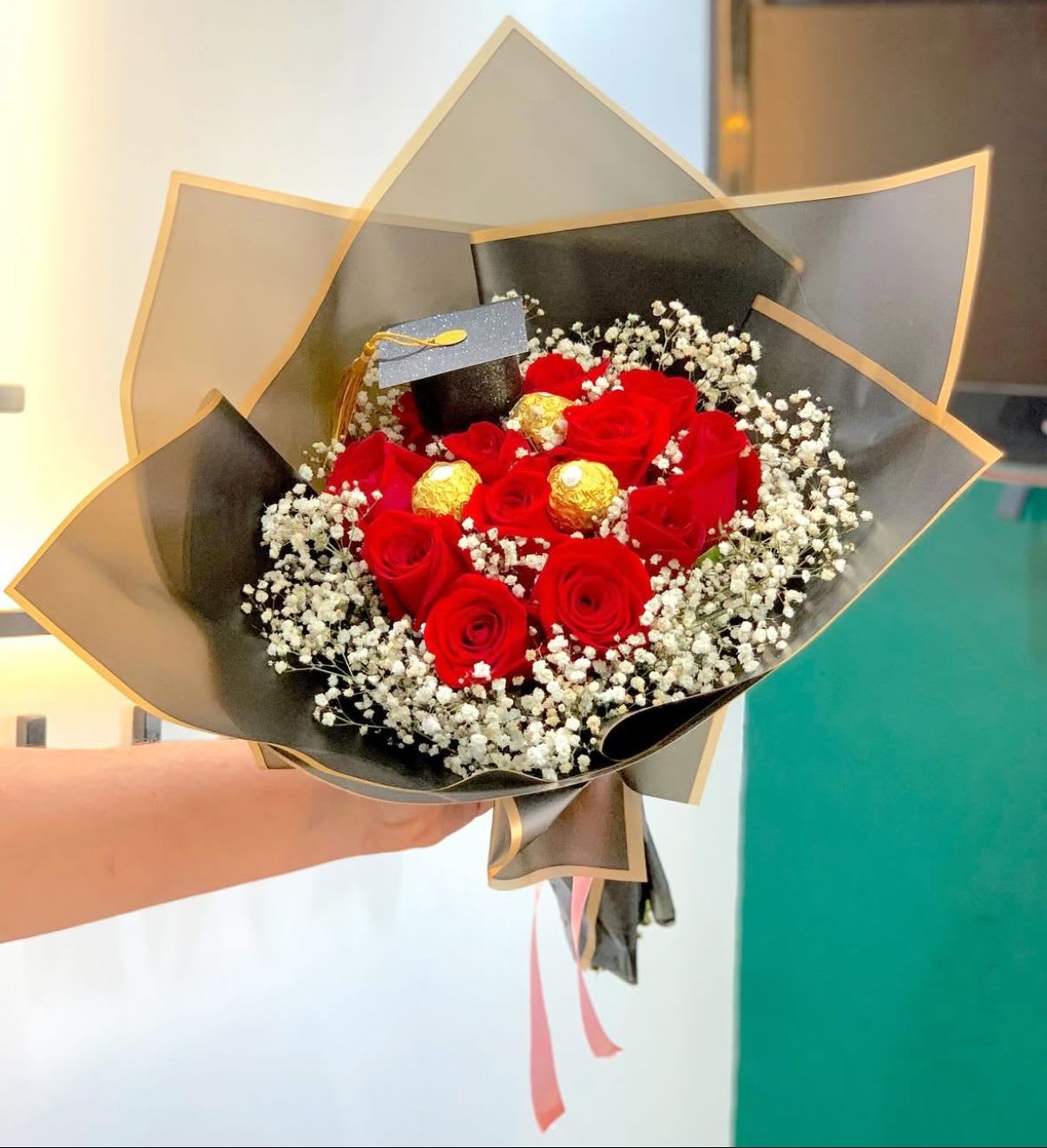 Golden Graduate Bouquet - Make their graduation day unforgettable with the Golden Graduate Bouquet OF 12 RED ROSES AND FERRERO ROCHER CHOCOLATES— a luxurious blend of classic romance and celebratory charm.   Wrapped in sleek black and gold-trimmed paper with a touch of pink ribbon, this bouquet is both sophisticated and celebratory — ideal for graduates stepping into a bright new chapter.  Whether for a friend, partner, or family member, this bouquet is more than flowers — it’s a proud moment wrapped in elegance and sweetness.