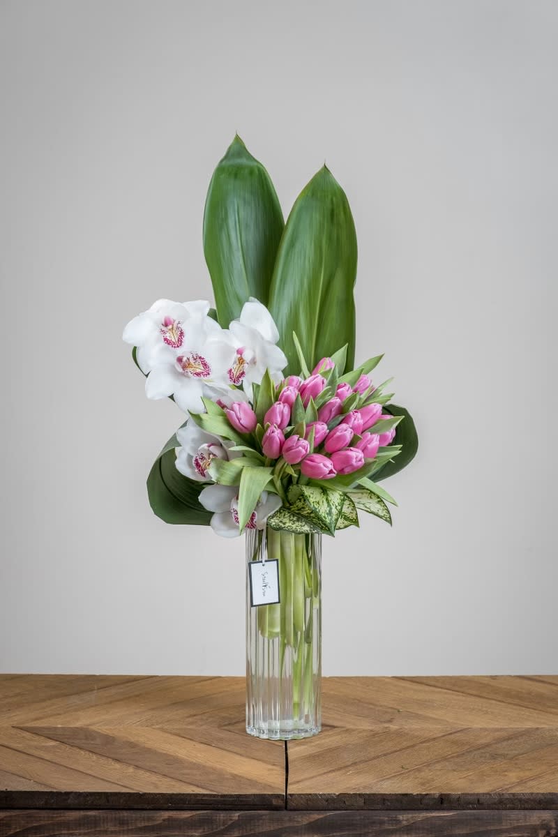 Jessica Jessica - A tall striped-textured glass cylinder towers high with stems of tropical, crisp white cymbidium orchids, bunches of flirty baby pink tulips, and complimented with gorgeous varieties of varying types of greenery. Show your flirtatious side with this impactful, romantic, and tall design this Valentine's Day. Measures approx. 30"H x 12"W