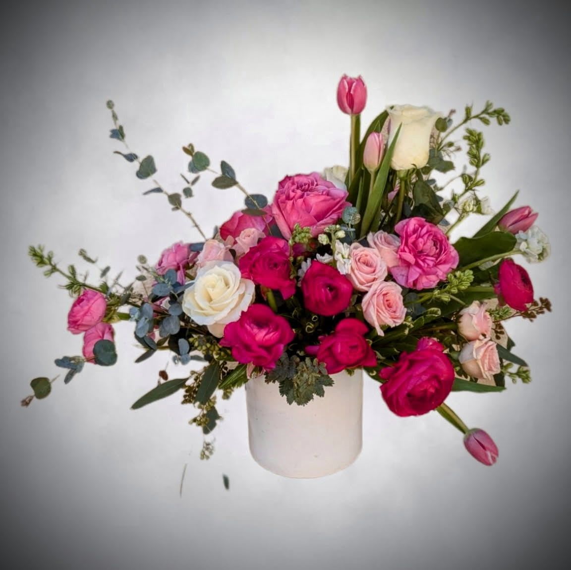 Blushing Bloomscape  - A tender composition of elegance and charm, Blushing Bloomscape features a delicate mix of magenta, blush pink, and creamy white roses, paired with romantic tulips and sprigs of eucalyptus. Artfully arranged in a matte white ceramic vase, this bouquet radiates gentle sophistication and heartfelt emotion.  Ideal for celebrating anniversaries, birthdays, or simply sending a warm gesture of love and admiration.    