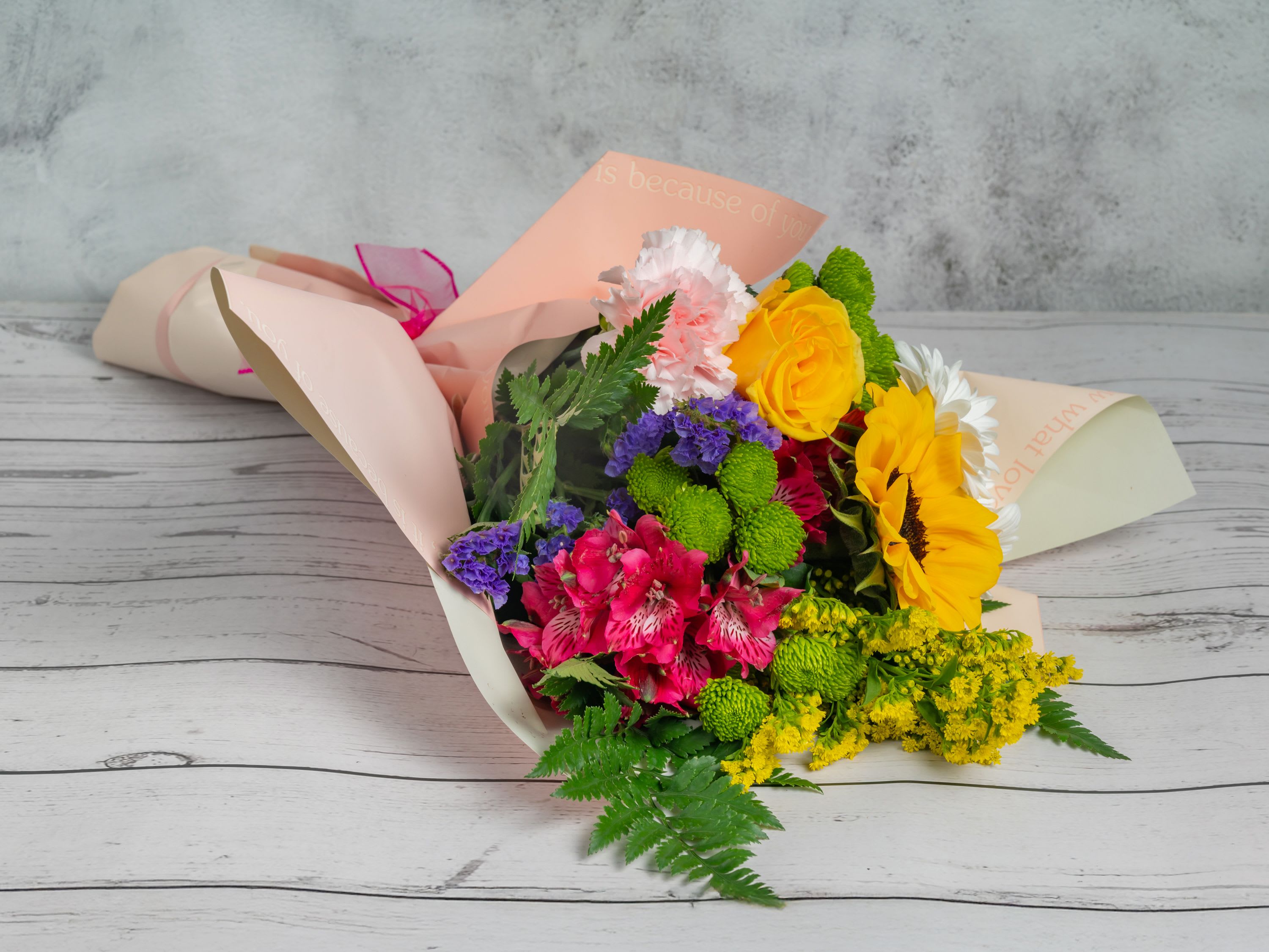Simple Life Simple Life - A vibrant and cheerful arrangement featuring sunny sunflowers, bright yellow roses, pink alstroemerias, white daisies, and playful green button poms, accented with purple statice and lush greenery. Wrapped in soft peach paper, this bouquet brings warmth and happiness to any occasion. Perfect for birthdays, congratulations, or simply to brighten someone’s day with its lively mix of colors and textures. Bring joy and simplicity with this charming bouquet.