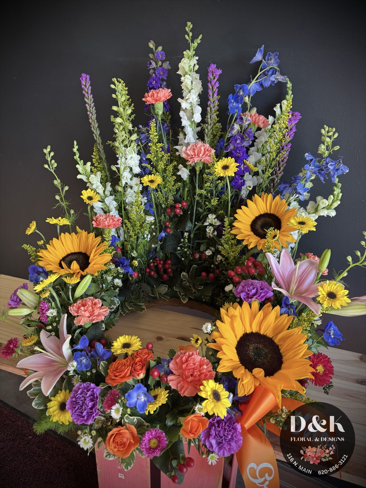 Sunflower wildflower urn arrangement - A beautiful urn arrangement with sunflowers and assorted colors of wildflowers. Colors and flowers may vary but the overall look and feel will remain the same. The urn is placed inside the wreath with the flowers framing your loved one. 
