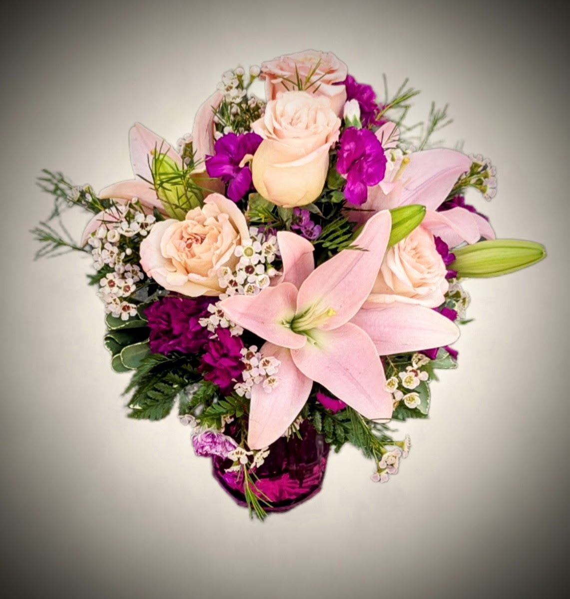 Blush & Bloom Elegance Bouquet Blush & Bloom Elegance Bouquet - Delicate and romantic, the Blush & Bloom Elegance Bouquet features soft pink lilies, creamy blush roses, and vibrant accents of purple and white blossoms. Designed in a chic jewel-toned vase, this arrangement exudes grace and charm, making it a beautiful gesture for anniversaries, love notes, or thoughtful surprises. Features: Elegant pink lilies for a soft, dramatic centerpiece Blush roses paired with rich purple blooms for contrast White waxflower and seasonal greenery for added texture Presented in a stylish deep pink glass vase Approximate size: 13" H x 10" W
