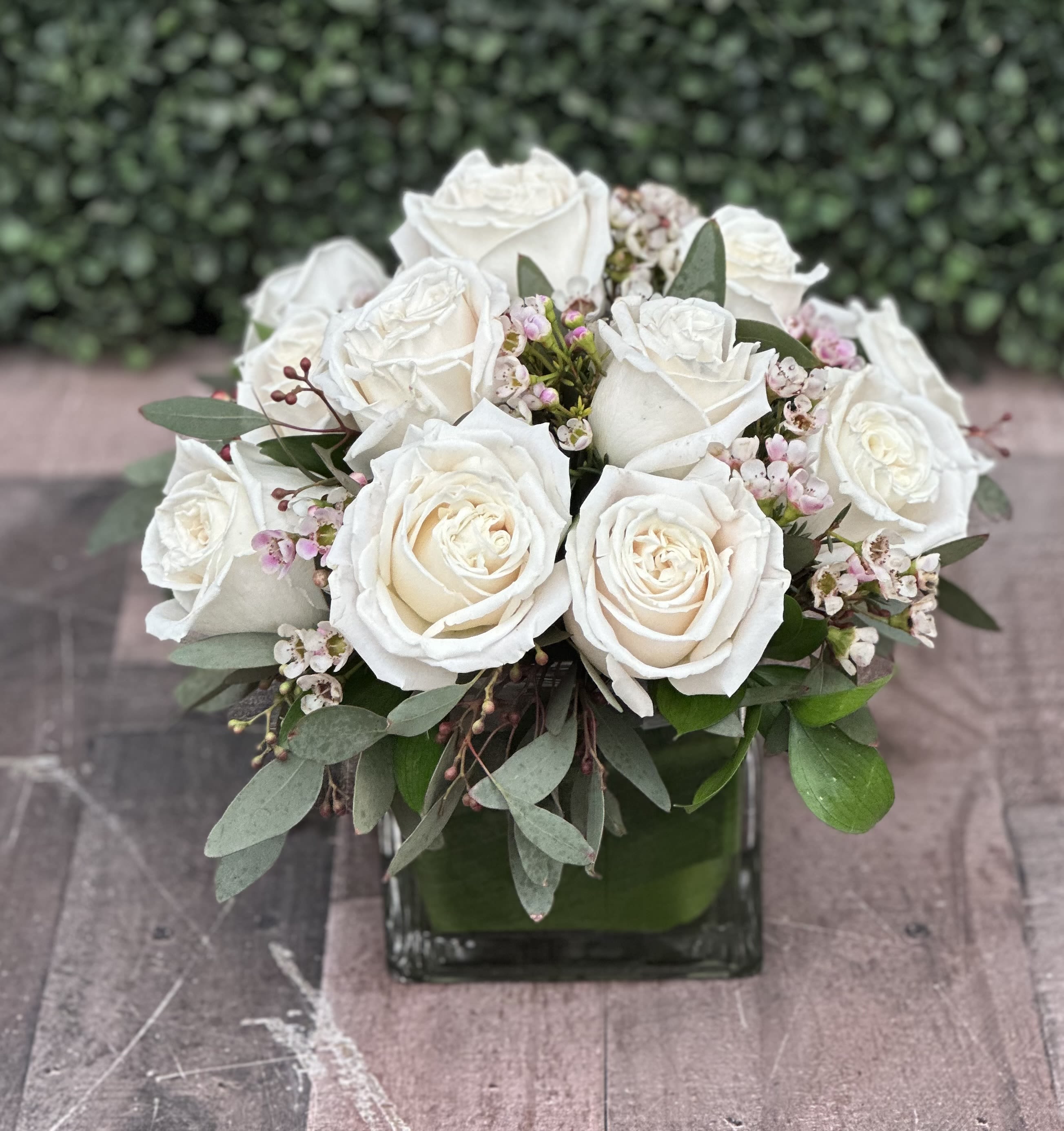 CLUSTER OF LOVE (WHITE) - This clustered arrangement comes designed with roses, seeded eucalyptus, wax flower and assorted greenery in a cube wrapped with a leaf.  