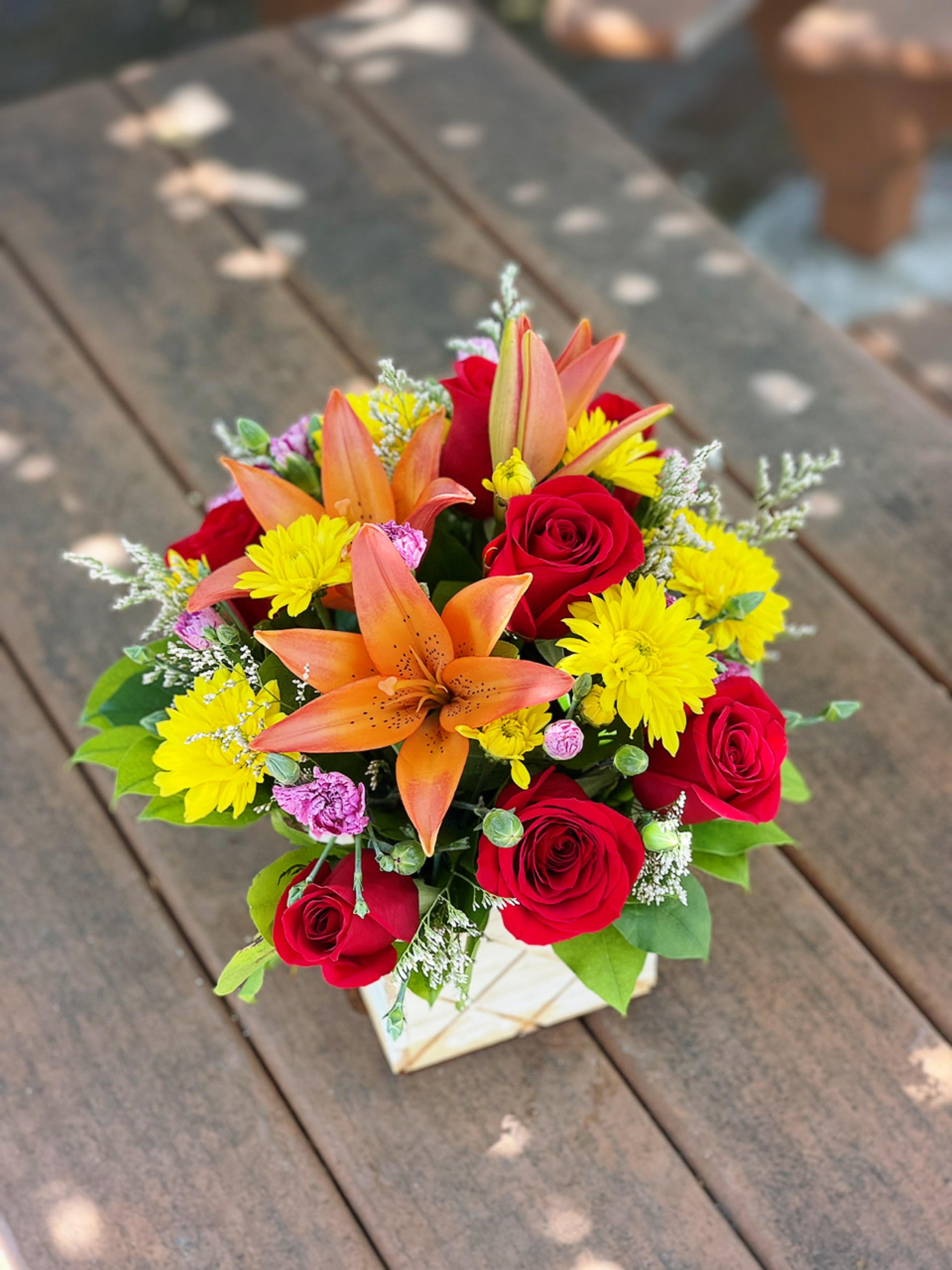 Bright Boxwood Bright Boxwood - - 2 Orange Lilies - 6 Red Roses - 3 Stems Yellow Cushions - 3 Stems Pink Carnations - Greenery and Fillers - 5 Inches Wooden Box