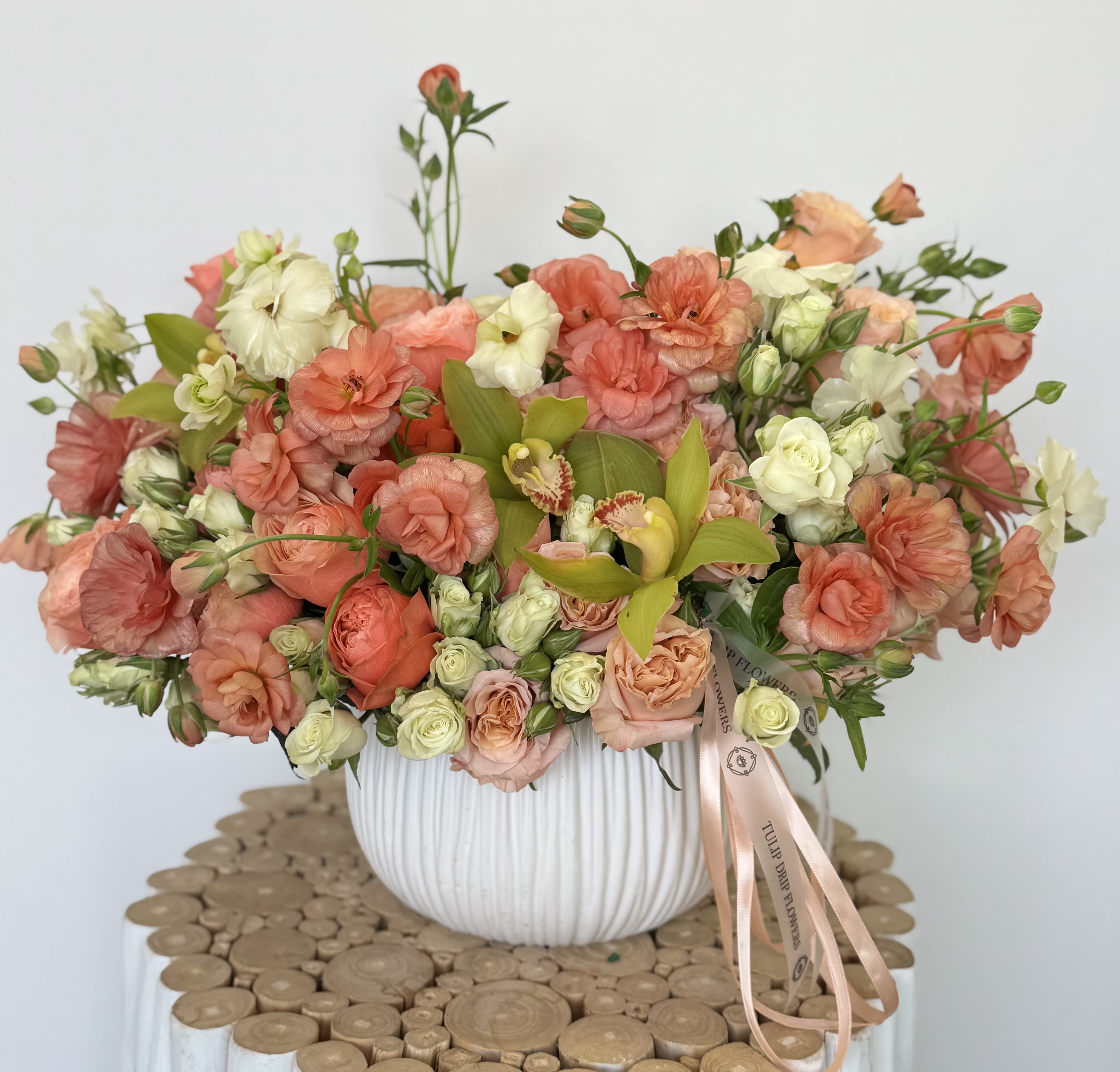 Coral Glow Garden Coral Glow Garden - Bright and cheerful, this garden-style arrangement features coral ranunculus, peach roses, cymbidium orchids, and lisianthus in a white ceramic bowl.