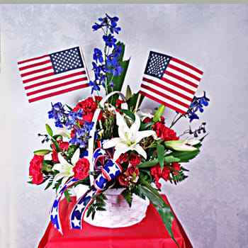 Old Glory - Traditional red, white and blue basket arrangement...patriotic; for celebrating a holiday or a person.  Blue delphinium, red carnations, white lilies, red spray roses - the foundation of the flowers...but certainly outshone by the American flags!  18H x 18W